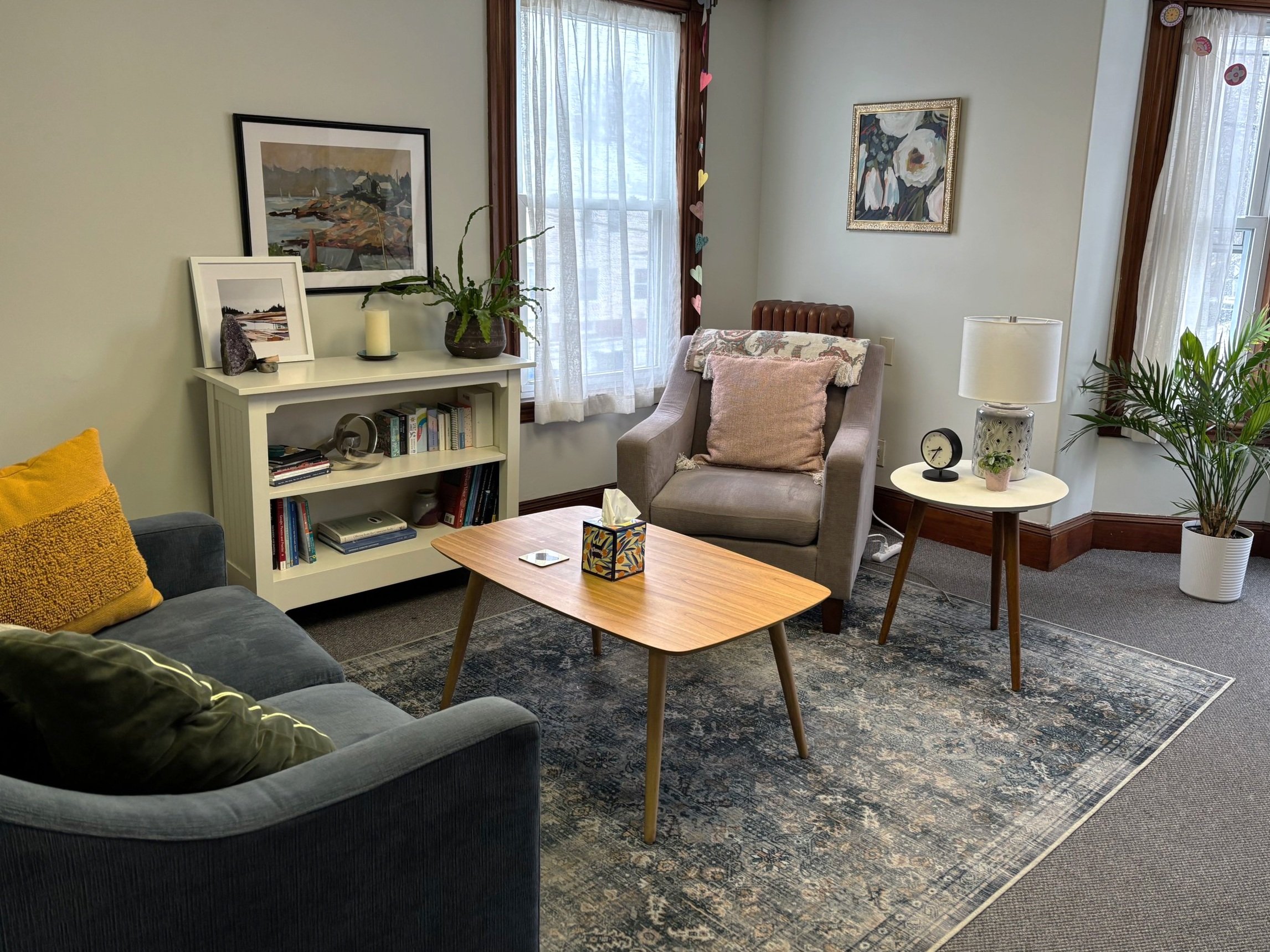 Living room with gray sofa, beige armchair, wooden coffee table, side table with lamp, window with white curtains, framed artwork, bookshelf, houseplant, and decorative pillows.
