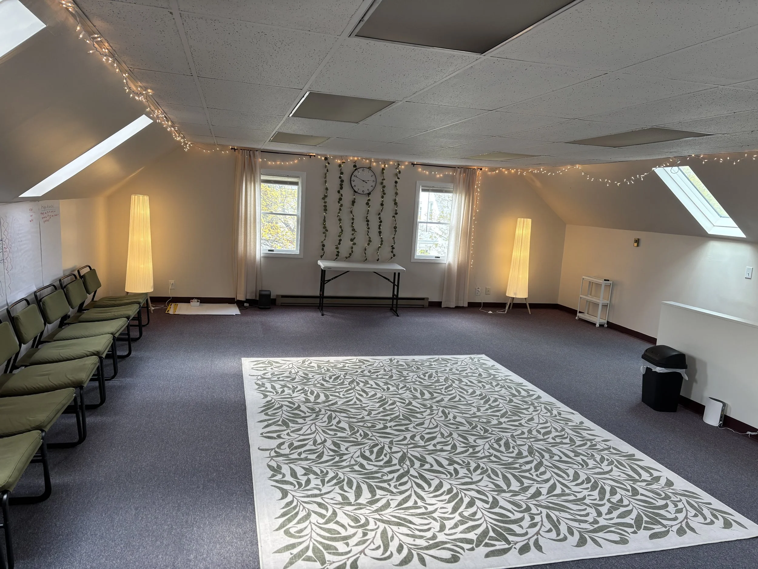 An empty room with a patterned rug, string lights, two tall floor lamps, chairs along the left wall, and windows providing natural light.