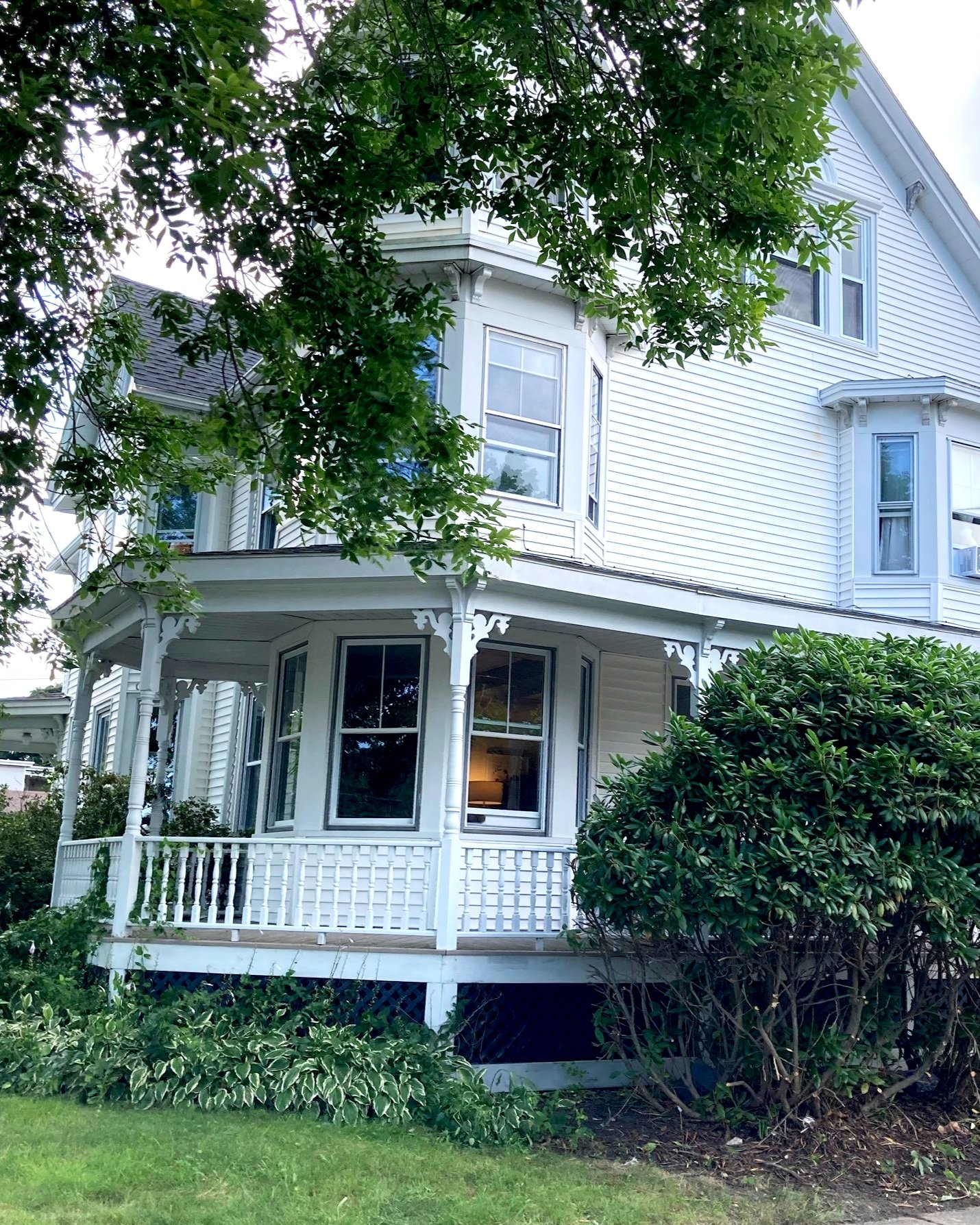 A white Victorian-style house with a front porch, surrounded by greenery including bushes and trees.