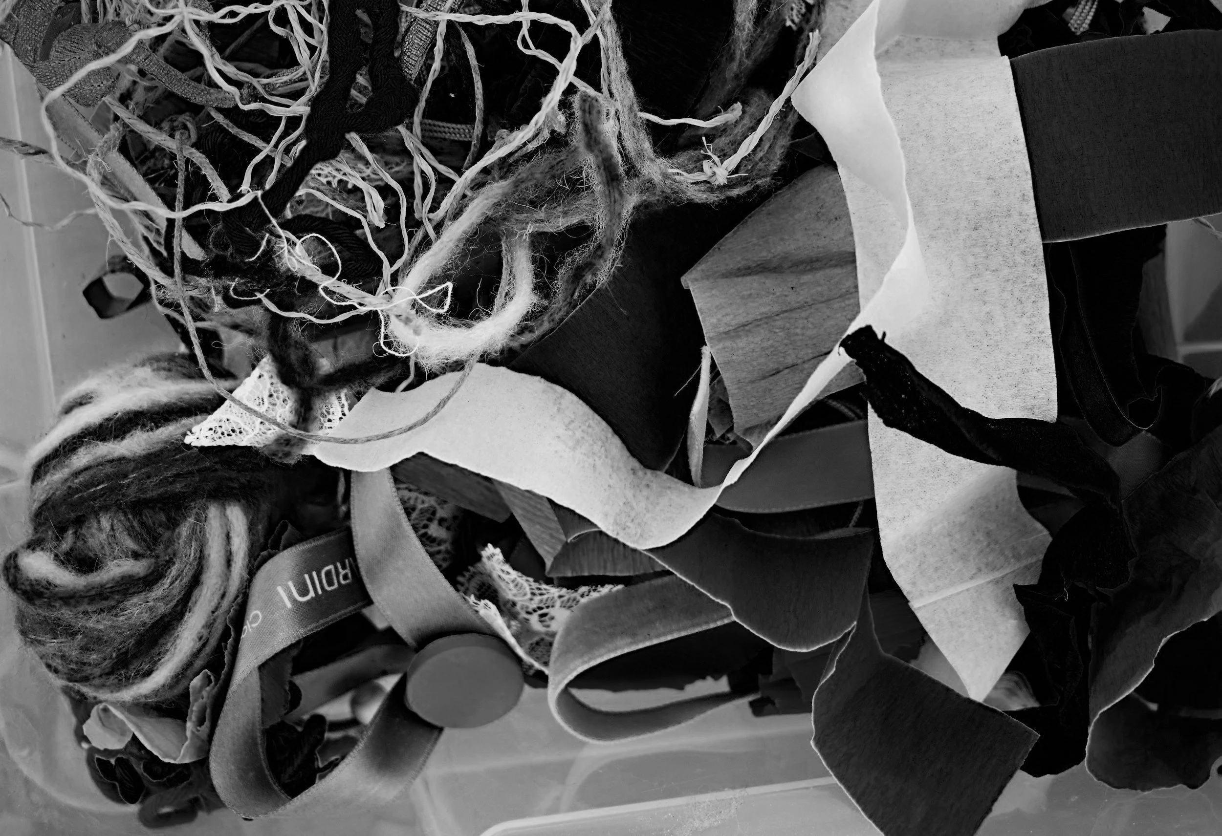 A tangled pile of various fabrics, textiles, and ribbons in grayscale, including yarn, lace, paper, and cloth scraps.