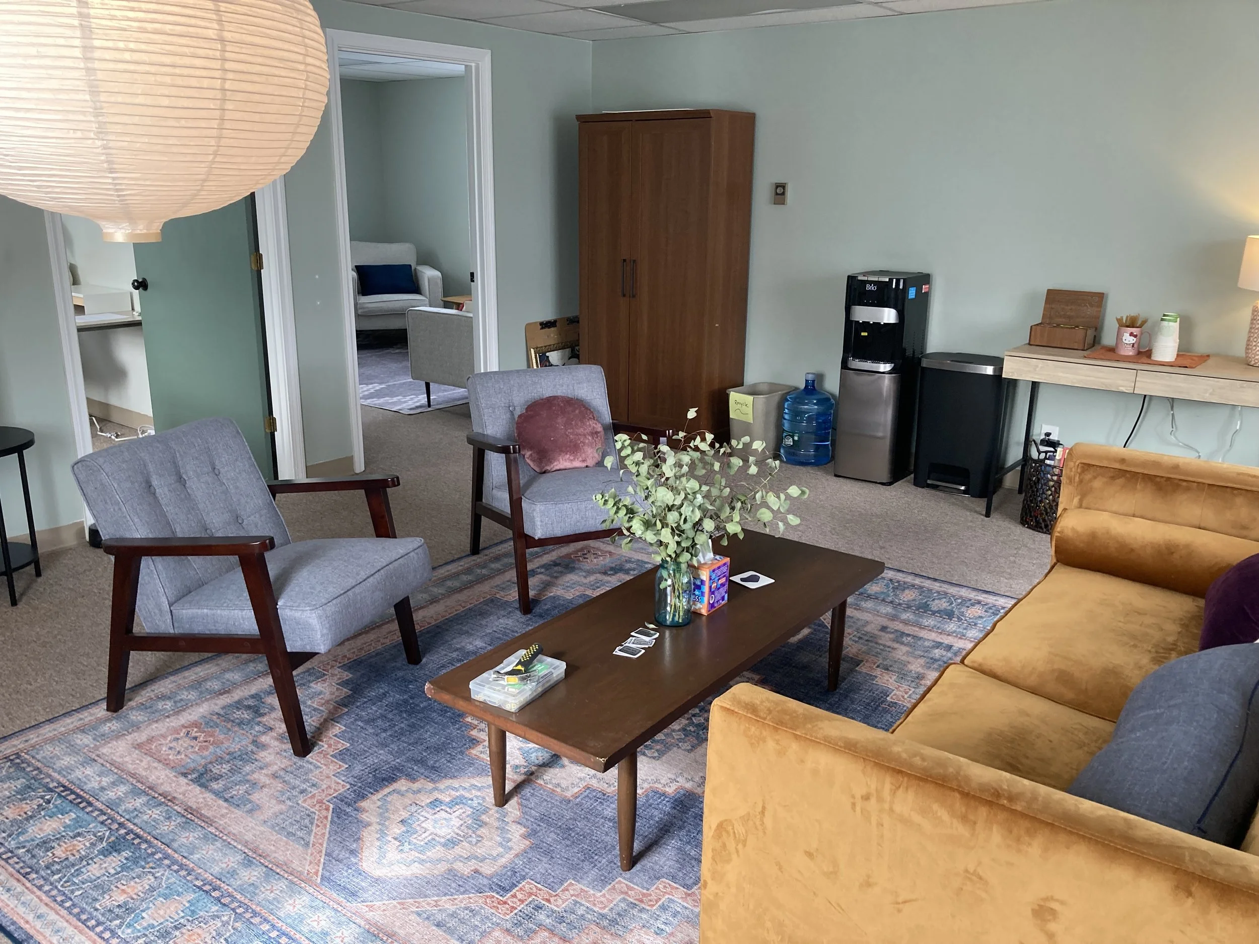 A cozy living room with a yellow velvet sofa, two gray armchairs, a wooden coffee table with a vase of greenery, and a patterned rug. In the background, there's a water cooler, a wooden cabinet, and a table with a lamp and supplies. Two doorways lead to other rooms.