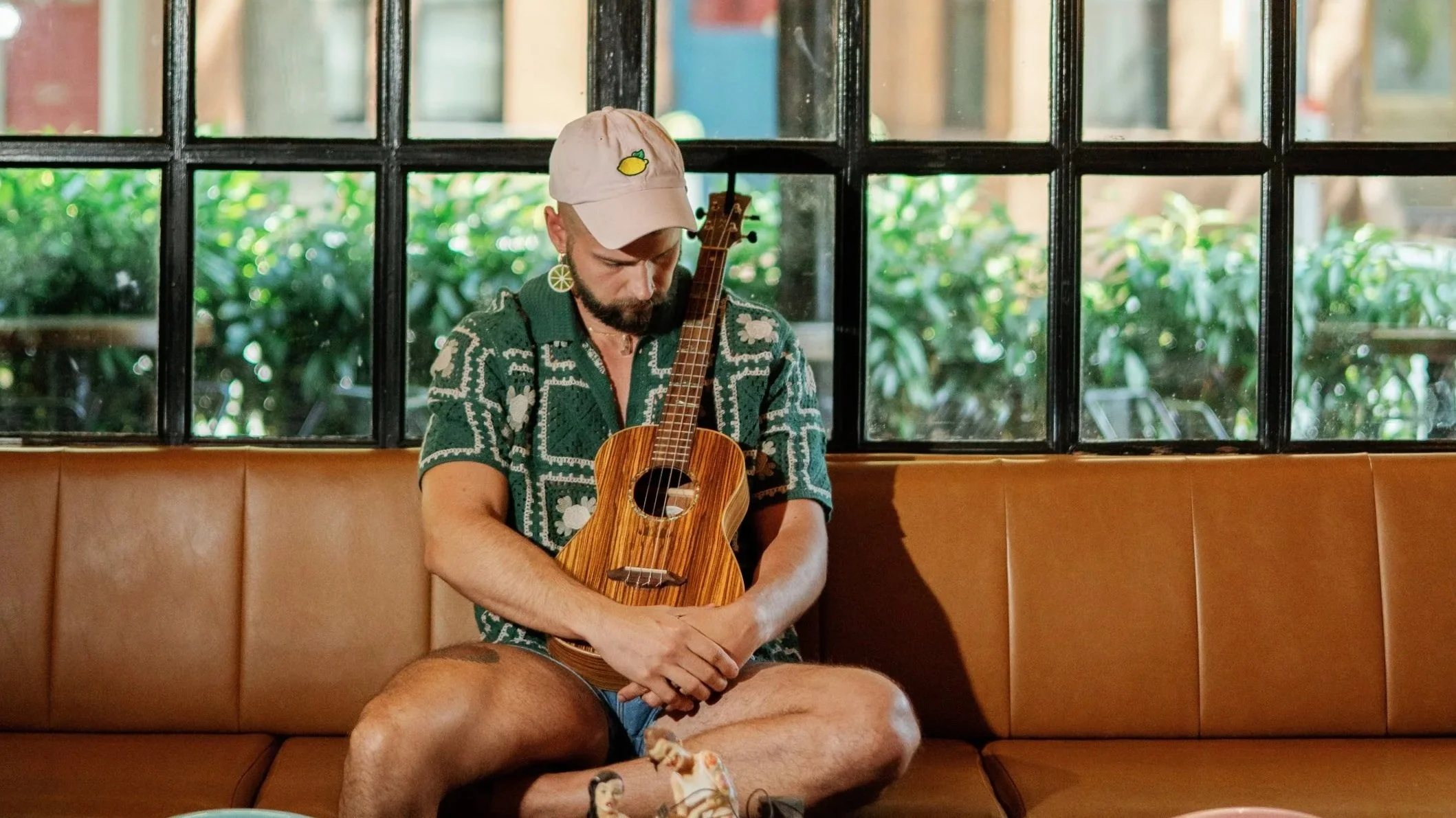 A man (Lemonboy) in a pink lemon hat and and a green crochet shirt sitting on a leather couch holding a ukulele.