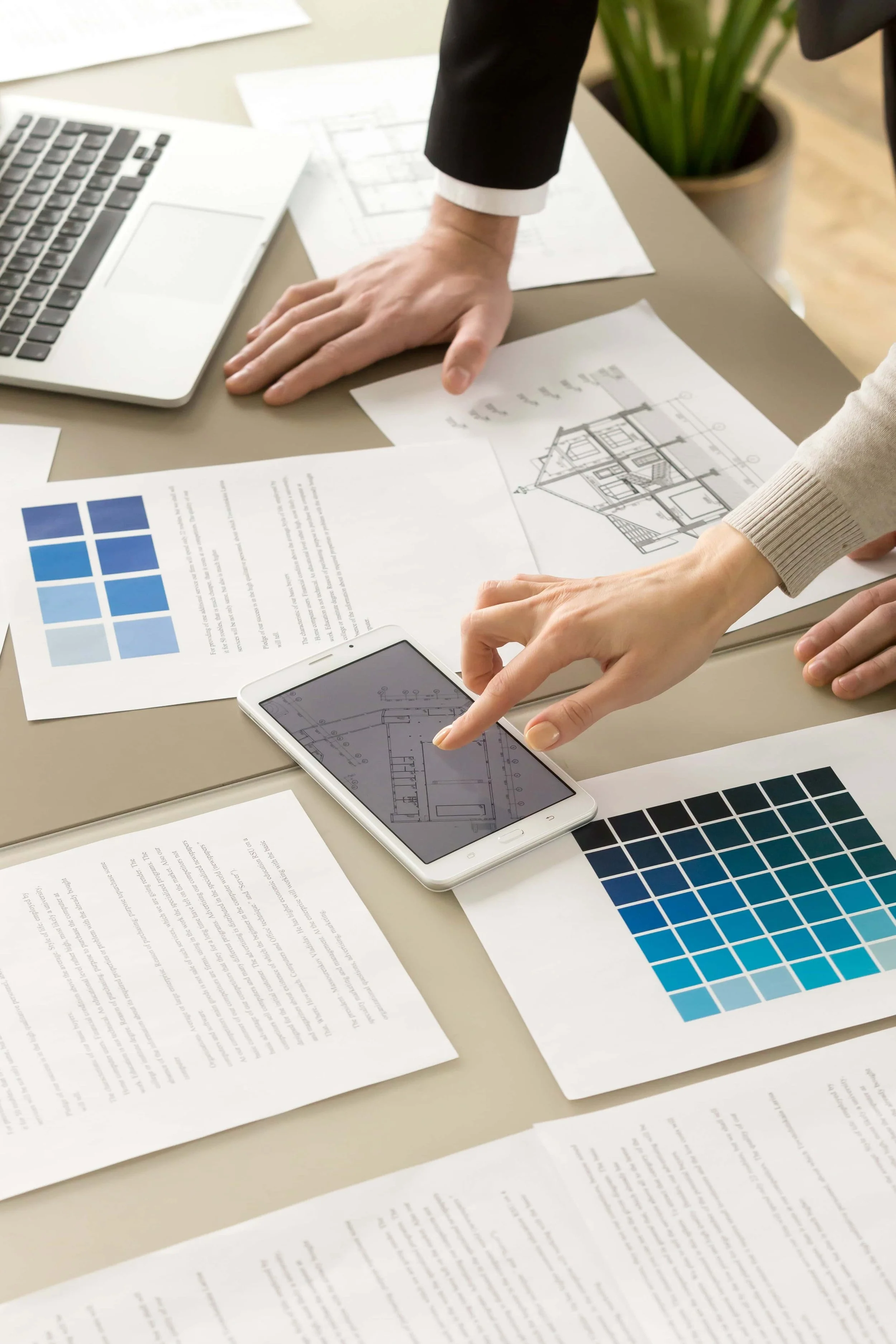 Two people discussing architectural plans, color swatches, and a laptop on a desk, with a potted plant in the background.