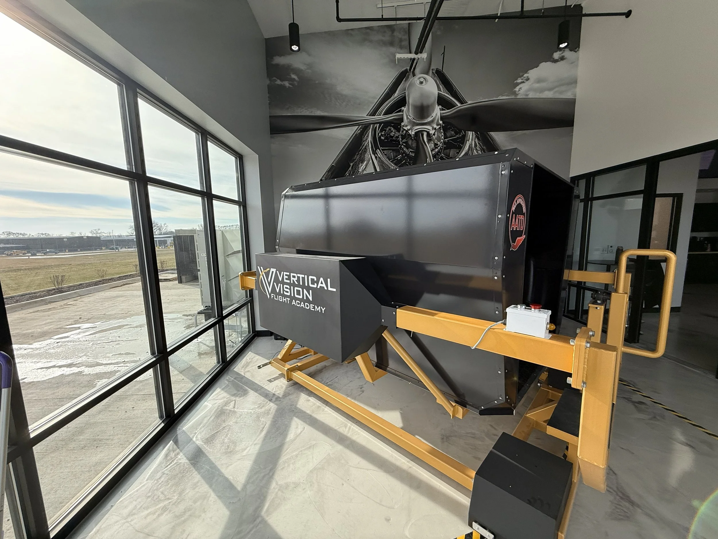 Indoor scene with a black helicopter engine mounted on a stand at the Vertical Vision Flight Academy, near large windows showing an outdoor tarmac area.
