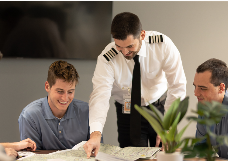 A pilot in uniform showing a map to two smiling passengers during a travel planning session.