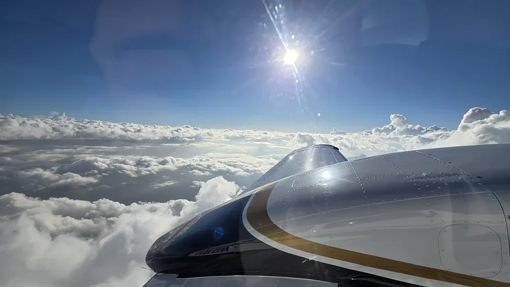 View from an airplane window showing parts of the aircraft and a sky filled with clouds and a bright sun.