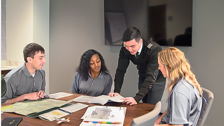 A diverse group of four people engaged in a discussion at a table, with a man standing and pointing at a book. The setting appears to be a meeting room with a large display on the wall.