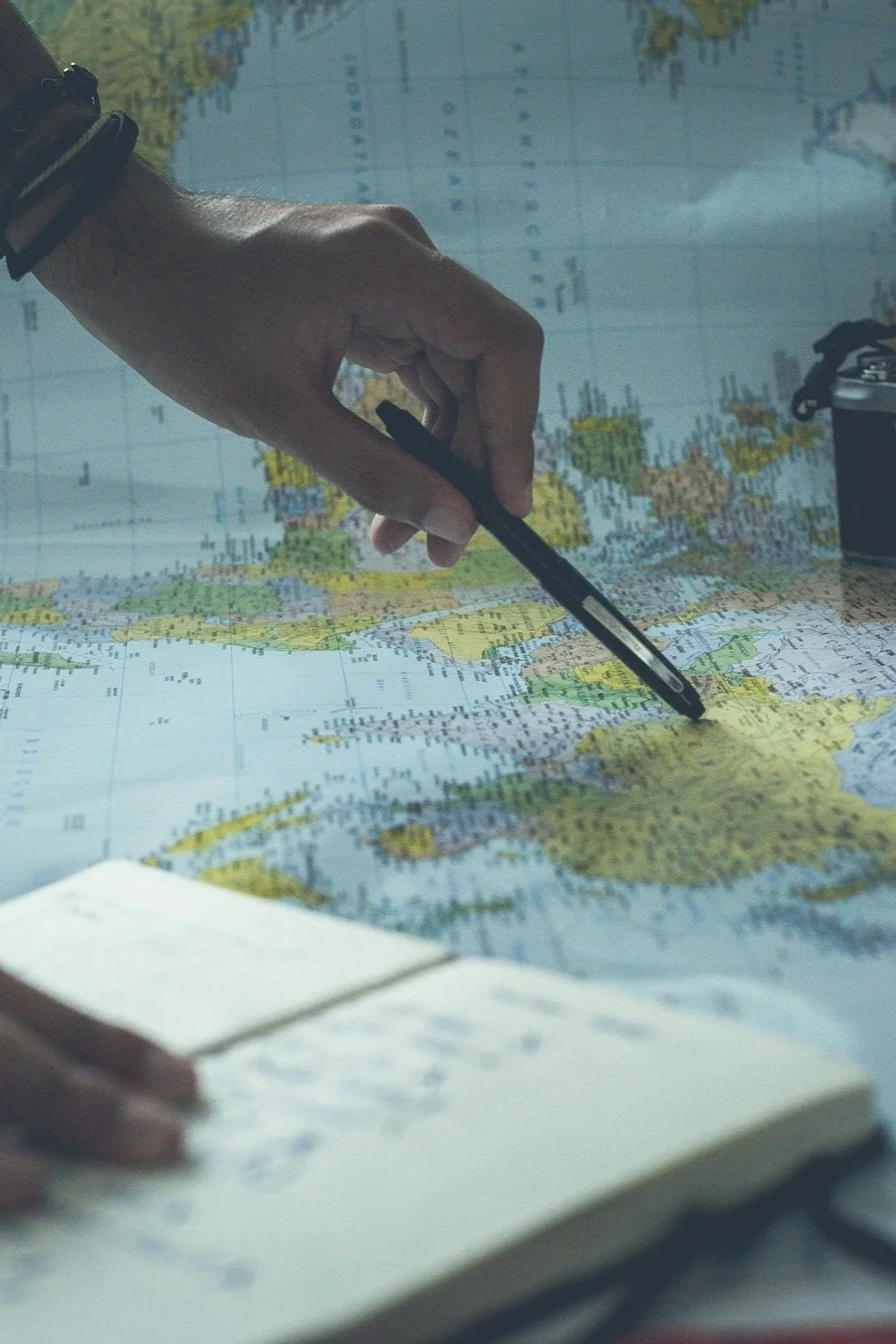 A person's hand pointing with a pen at a world map spread out on a table.