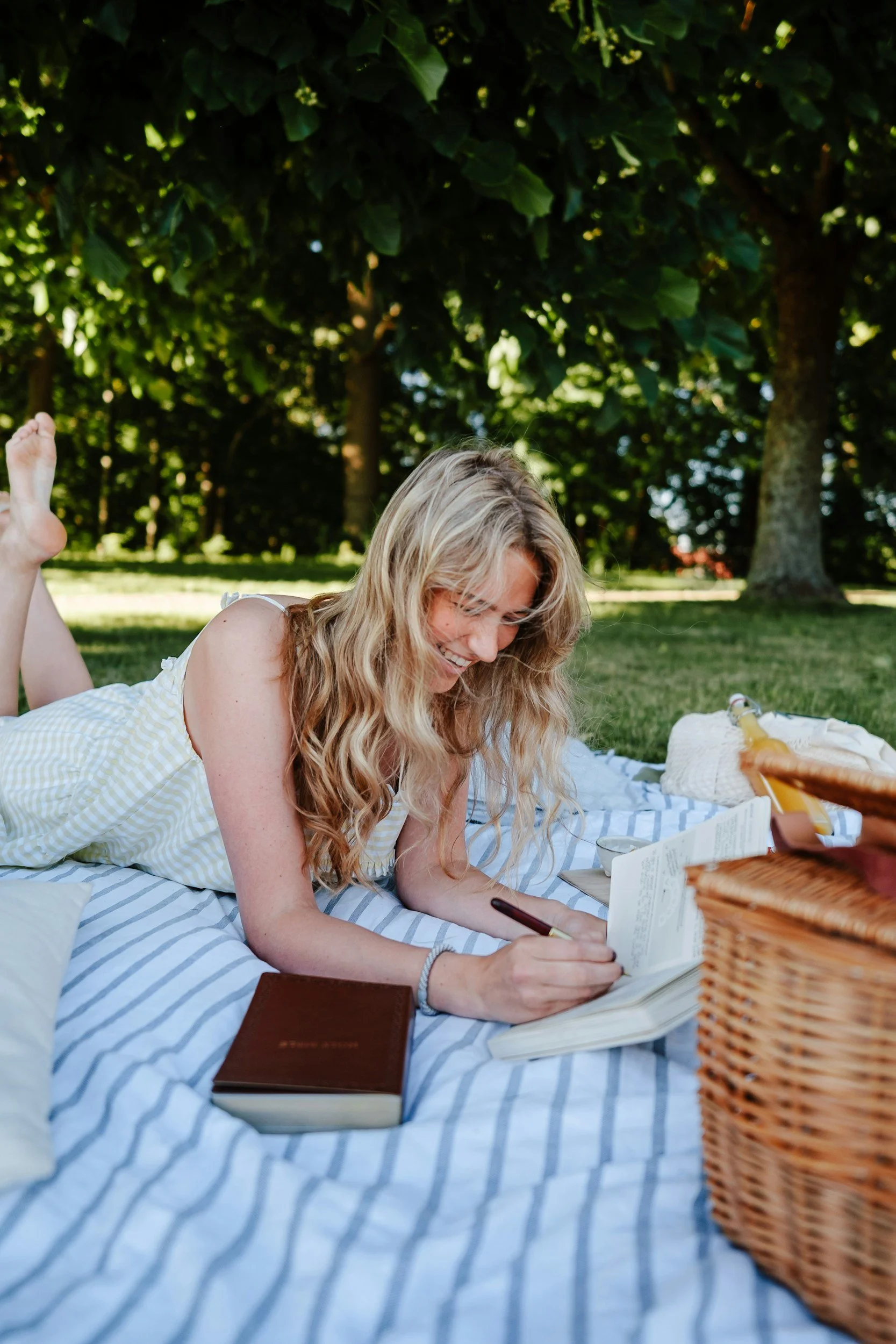 A woman with long blonde hair lying on her stomach on a blue and white striped blanket outdoors, writing in a notebook. There are books, a basket, and a glass bottle nearby, with green trees in the background and sunlight filtering through the leaves.