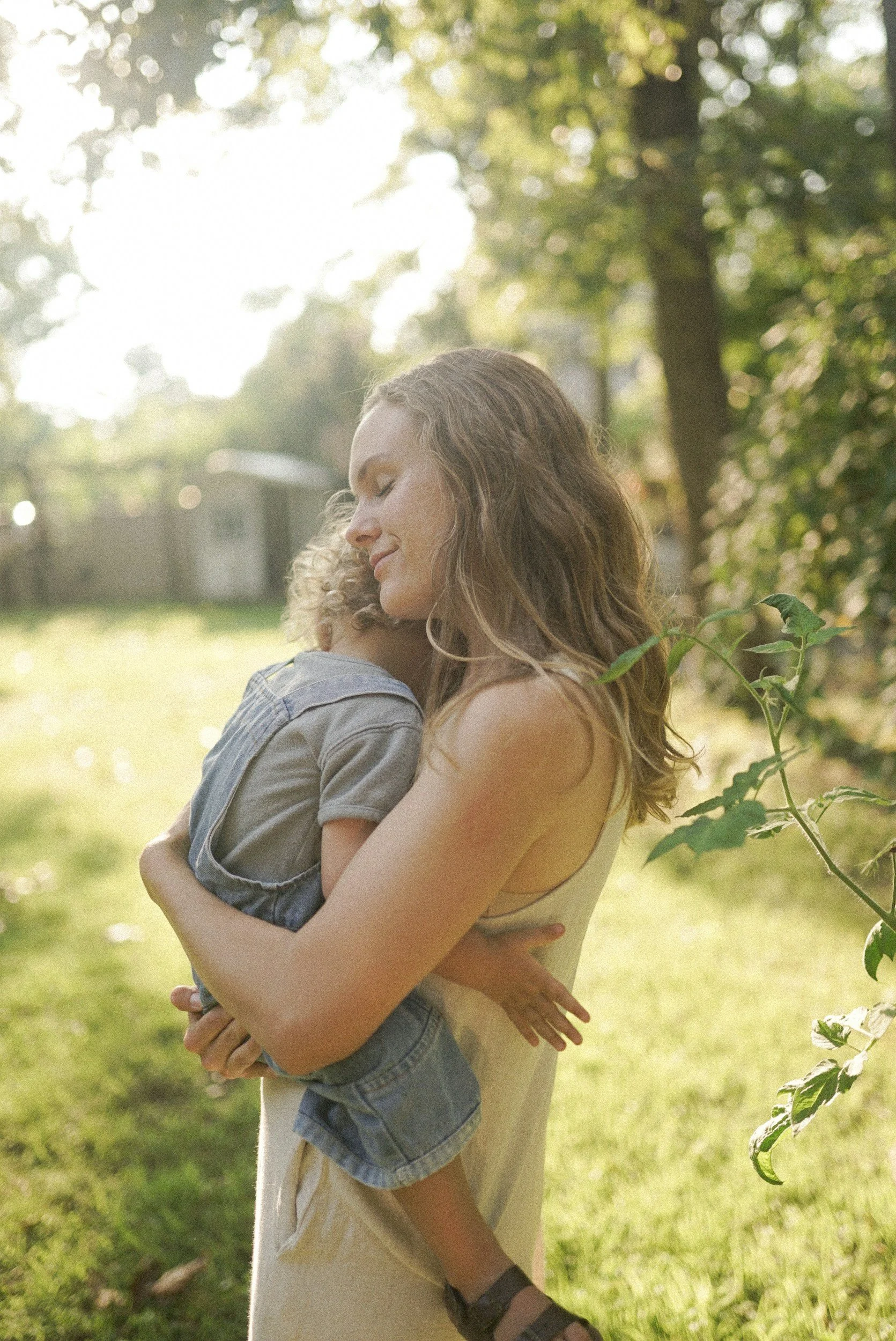 A woman holding a young child outdoors in a park during daytime, with sunlight filtering through trees in the background.