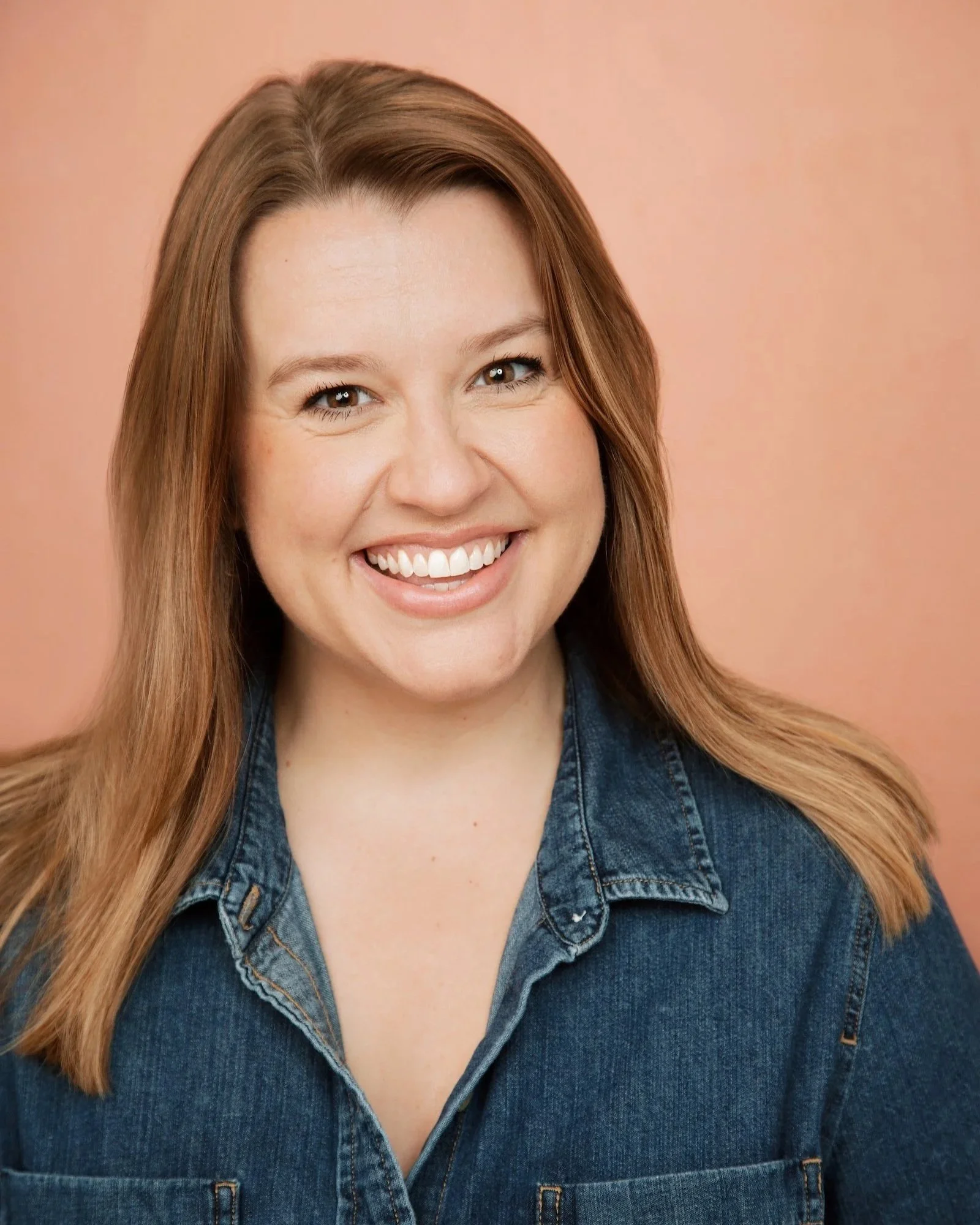 A woman with shoulder-length light brown hair smiling, wearing a denim shirt, against a peach-colored background.