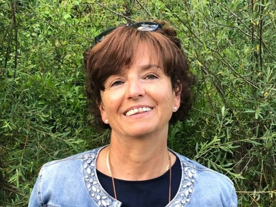 Eine lachende Frau mit kurzen braunen Haaren, Sonnenbrille auf dem Kopf, trägt eine schwarze Top, eine Jeansjacke und steht vor grünen Büschen.