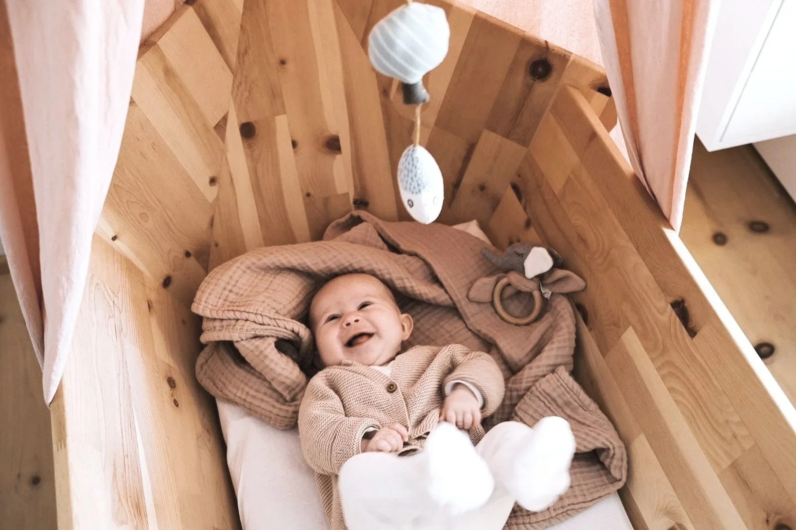 Lächelndes Baby, liegend in einer Babywiege aus Zirbe, mit Stofftieren und einem Mobile darüber.