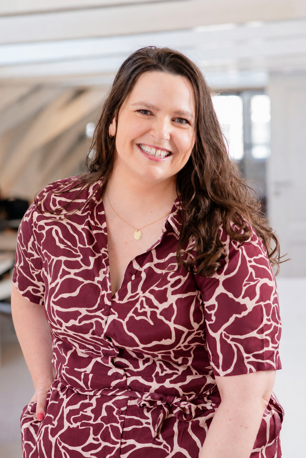 A woman with long curly brown hair smiling and wearing a maroon and white patterned dress in an indoor setting with natural light.