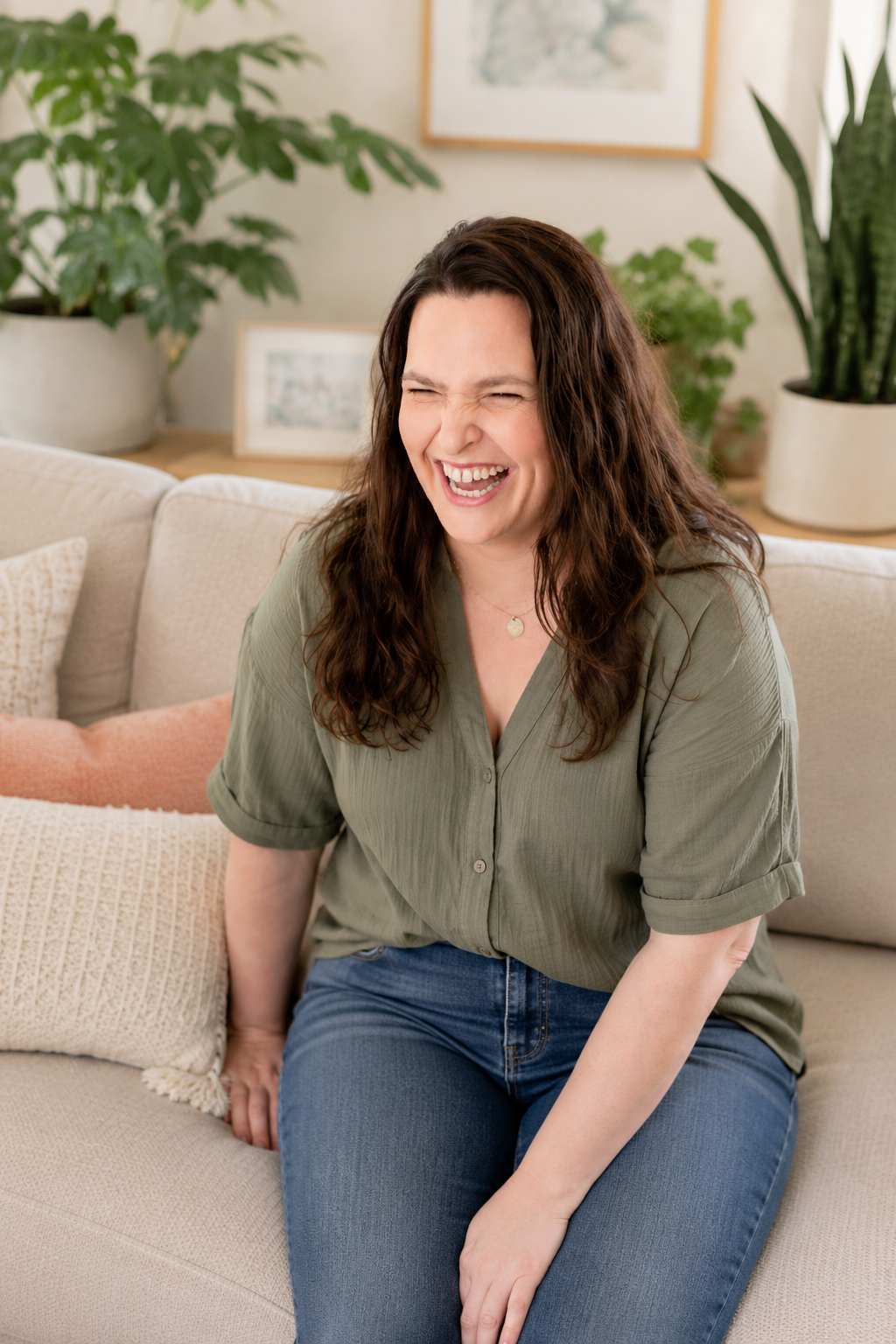 A woman with dark brown, curly hair laughing and sitting on a beige sofa with patterned pillows in a living room with green plants and art on the wall.