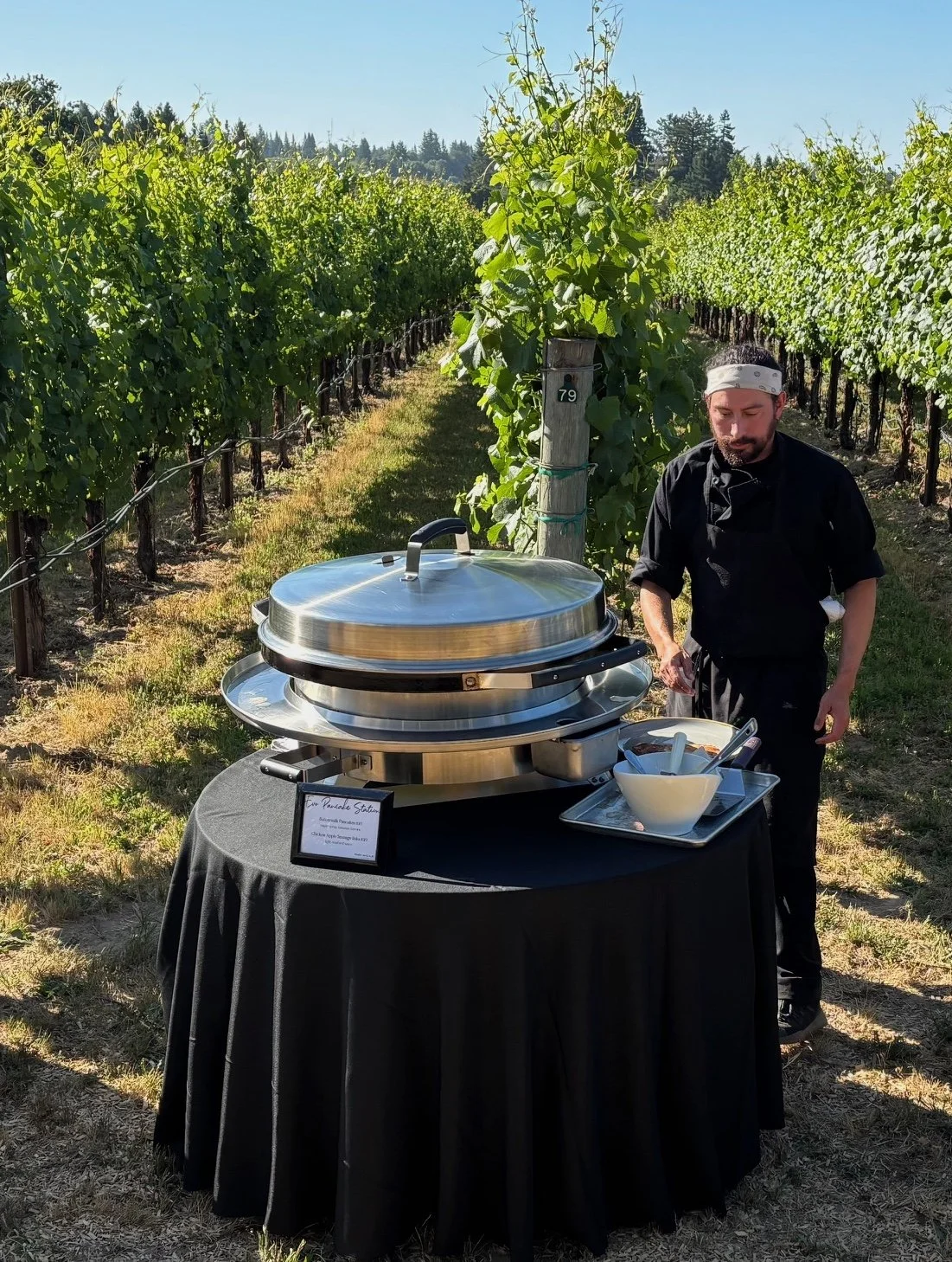 A man in black clothing with a white headband stands behind a round table with a black tablecloth. On the table, there is a chafing dish and a large white bowl with serving utensils. The setting is outdoors in a vineyard with rows of grapevines under a clear blue sky.