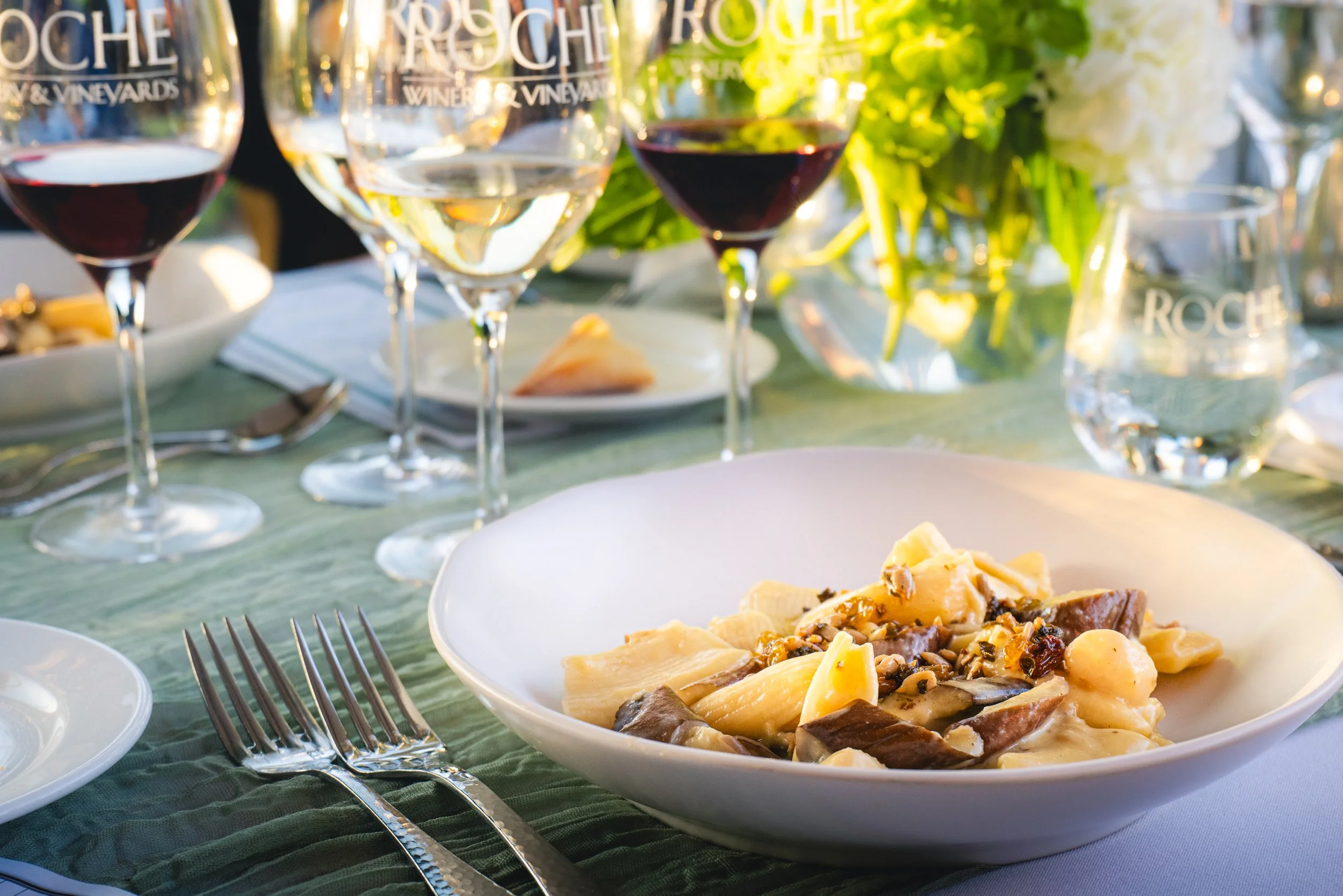 A table setting with four glasses of wine, a bowl of pasta with sauce, and a glass pitcher of water in the background. The table has a green tablecloth, and there is a flower arrangement visible behind the glasses.