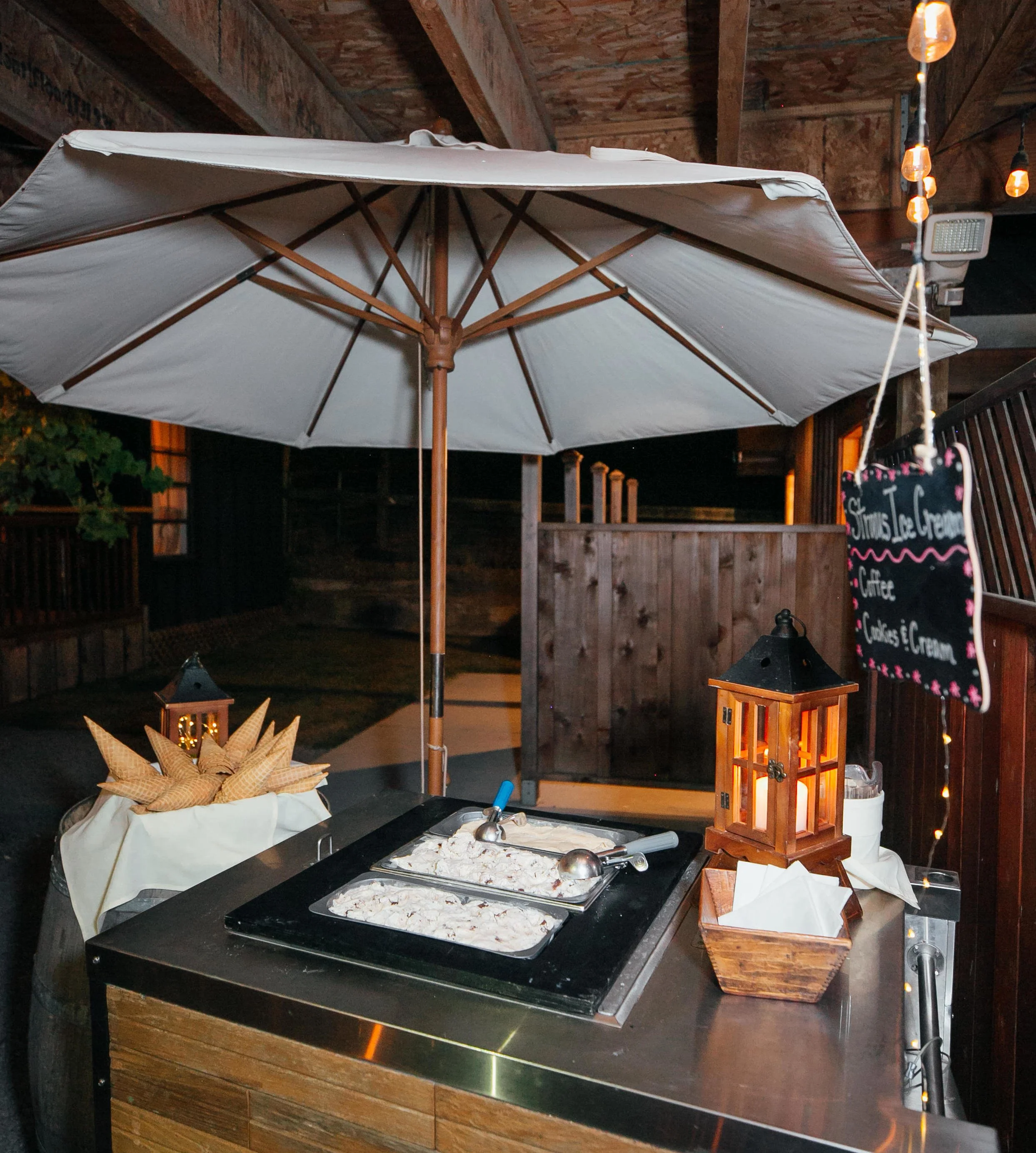 Ice cream serving station with two trays of ice cream, a scoop, a large umbrella, decorative lanterns, and a chalkboard sign for toppings, set up outdoors at night.