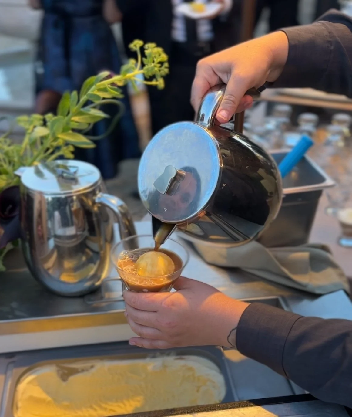 Enjoy a delicious dessert as coffee is poured over vanilla ice cream in a glass cup, set against a backdrop of ice cream containers, all brought to you by Park Avenue Catering.