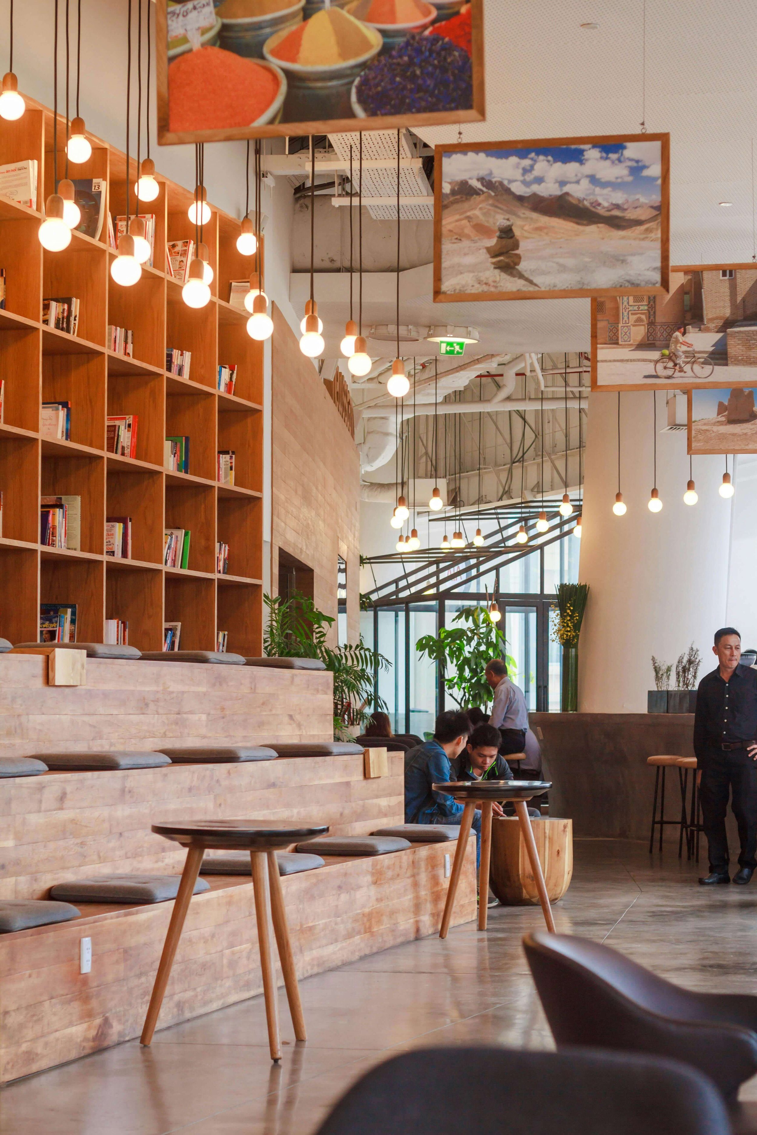Intérieur d'un café moderne avec bibliothèque en bois, luminaires suspendus, plantes vertes, et quelques clients assis et discutant.