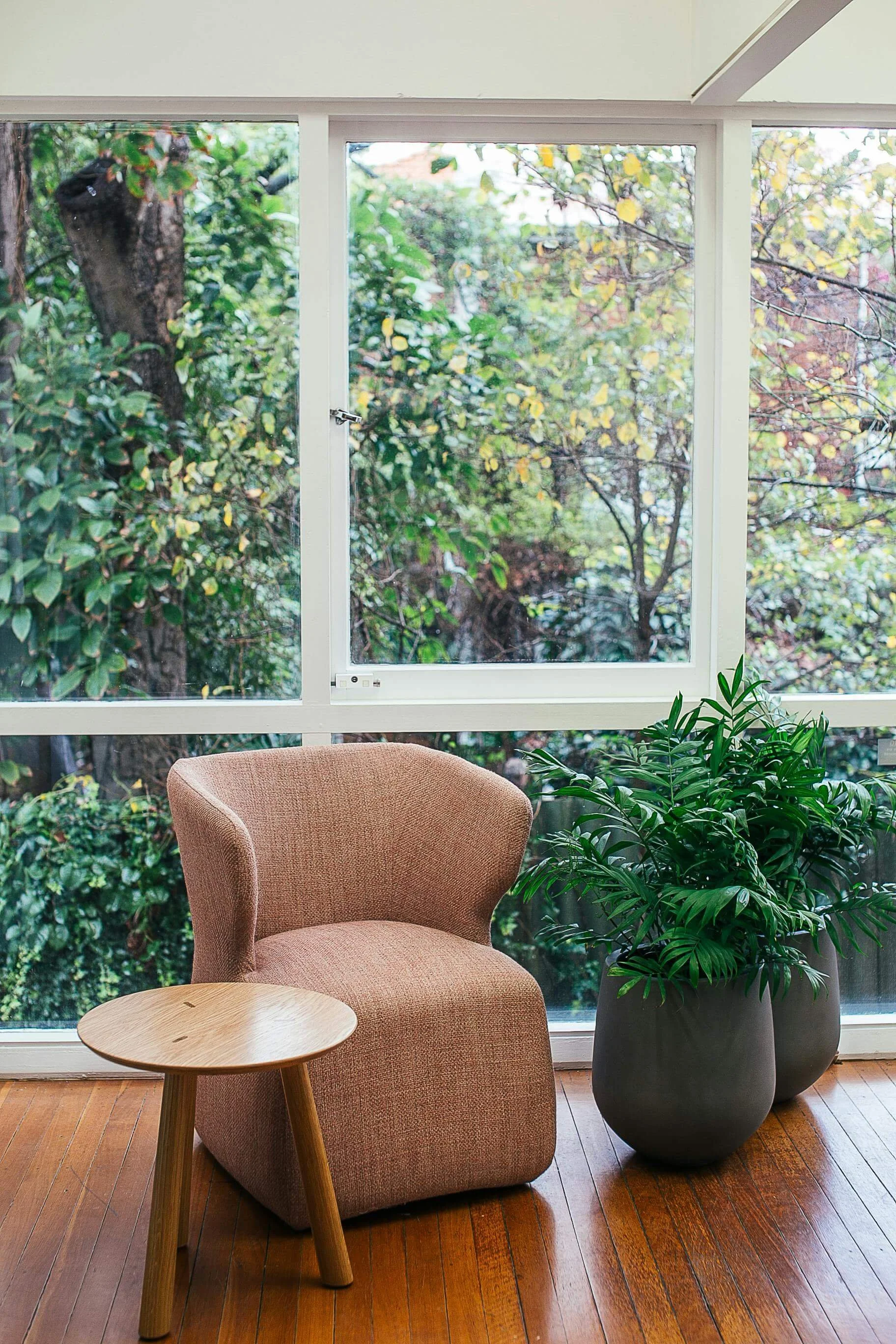 Salon intérieur avec un fauteuil rose en tissu, une petite table en bois, deux grands pots avec des plantes vertes, grande fenêtre donnant sur un jardin avec des arbres.