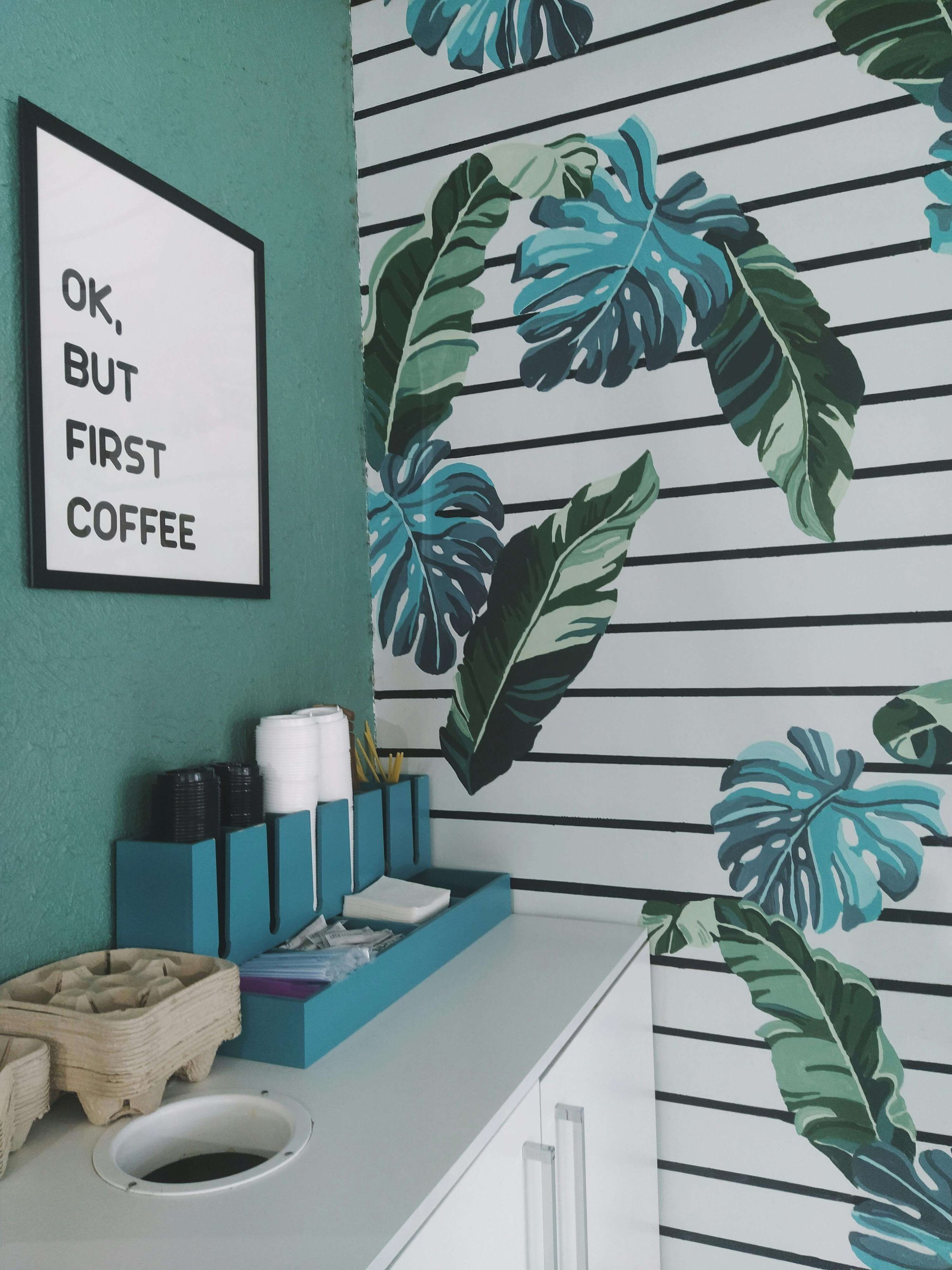 Mur avec un mur de plantes tropicales et un panneau blanc avec texte noir : « OK, BUT FIRST COFFEE ». Un comptoir blanc avec une boîte bleue contenant des gobelets en plastique noir et blanc, ainsi que des accessoires en plastique, et un panier en os