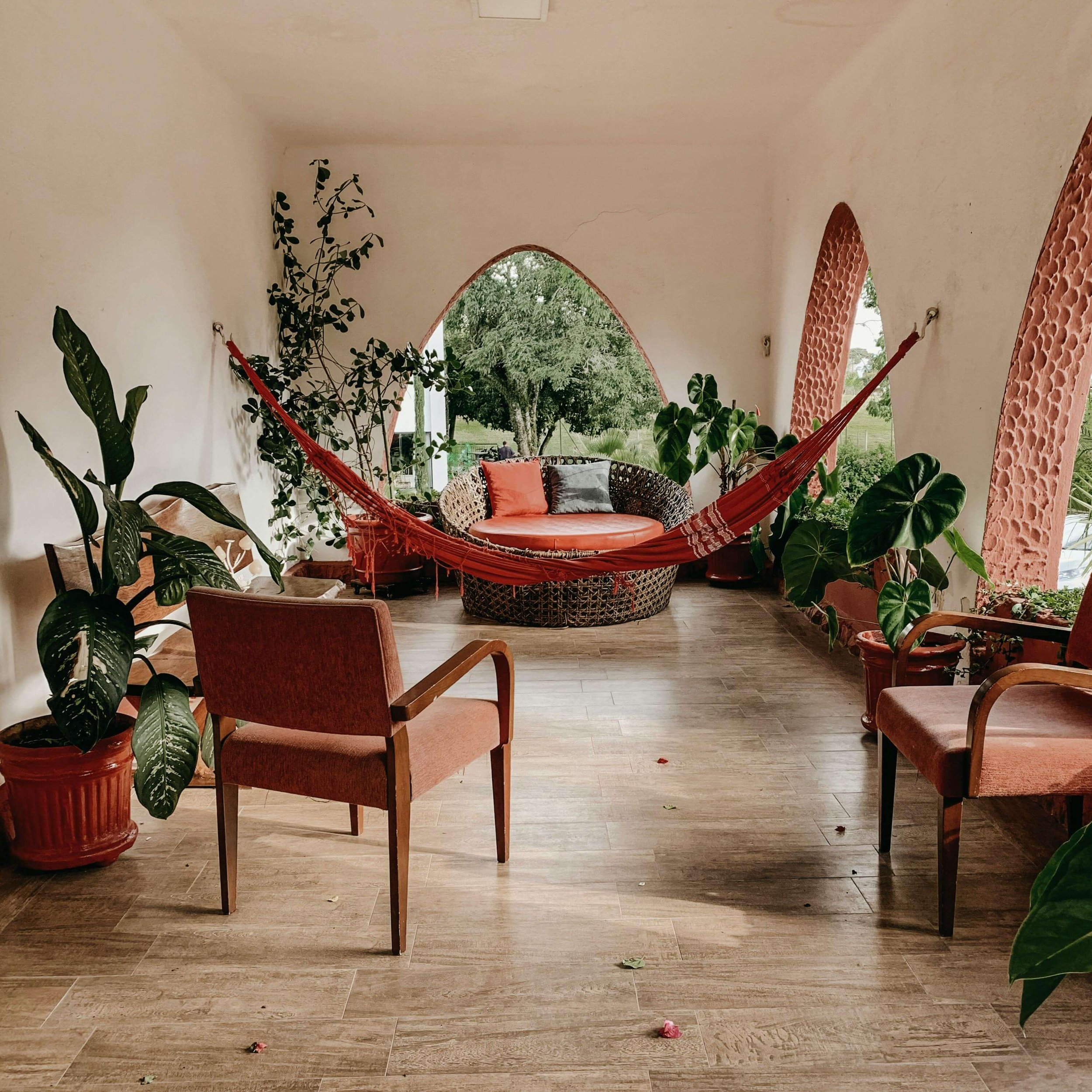 Salon avec une balançoire rouge, fauteuils en bois avec tissu rouge, plantes vertes en pot, fenêtres arquées view sur jardin