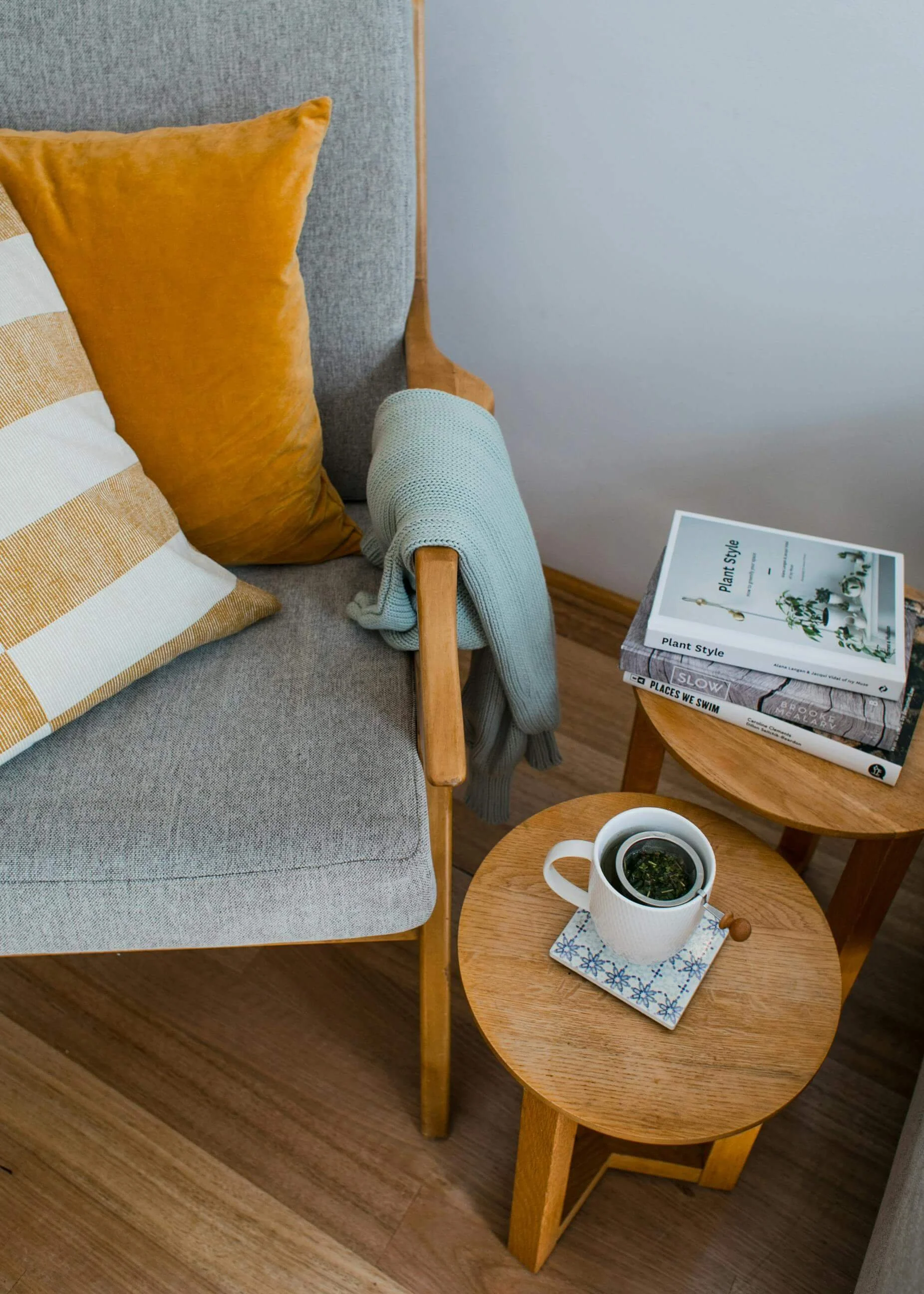 Une table en bois avec une tasse de thé et un miroir, une étagère en bois avec des livres et une banquette en tissu gris avec des coussins jaunes et un plaid gris.