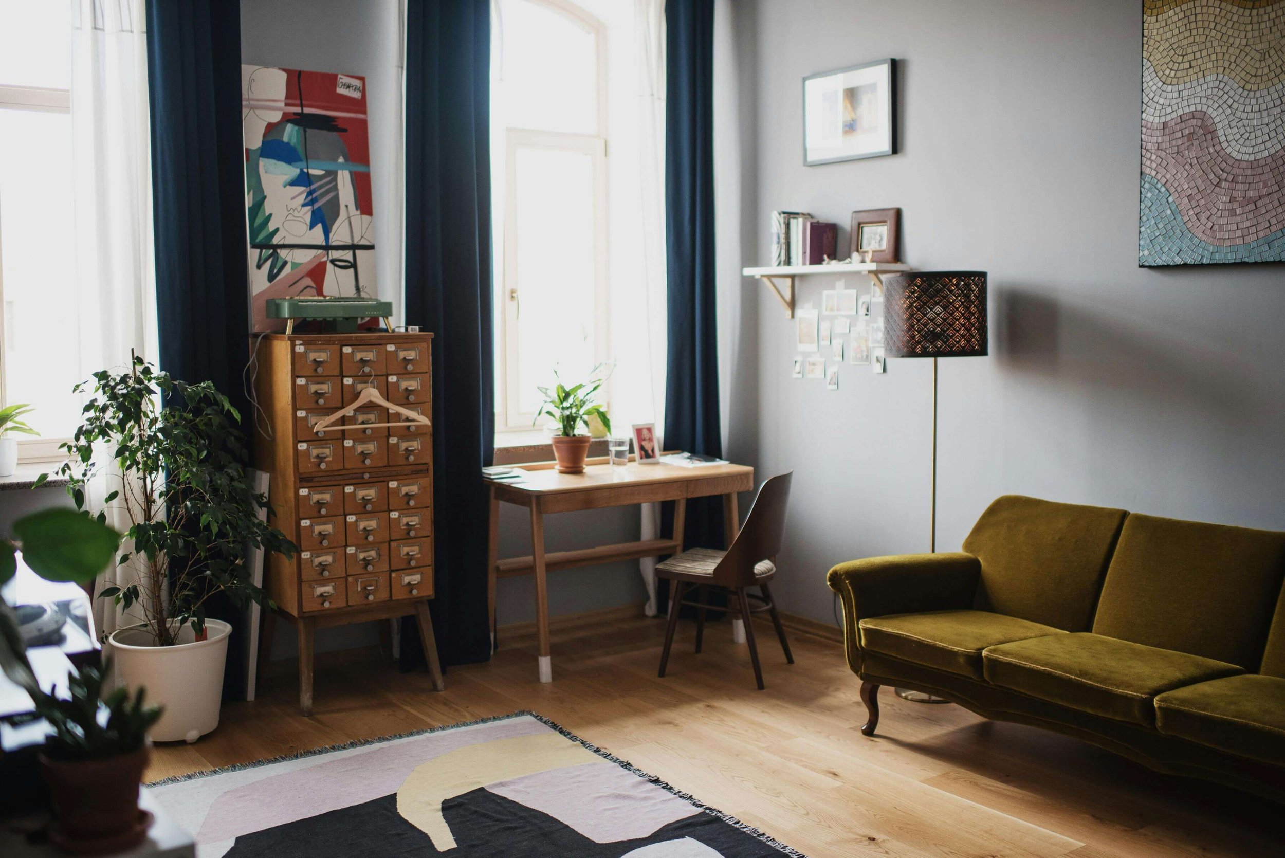 Salon lumineux avec canapé vert, bureau en bois, étagère avec livres, lampe à pied, œuvres d'art murales, plantes d'intérieur, tapis moderne sur parquet en bois.