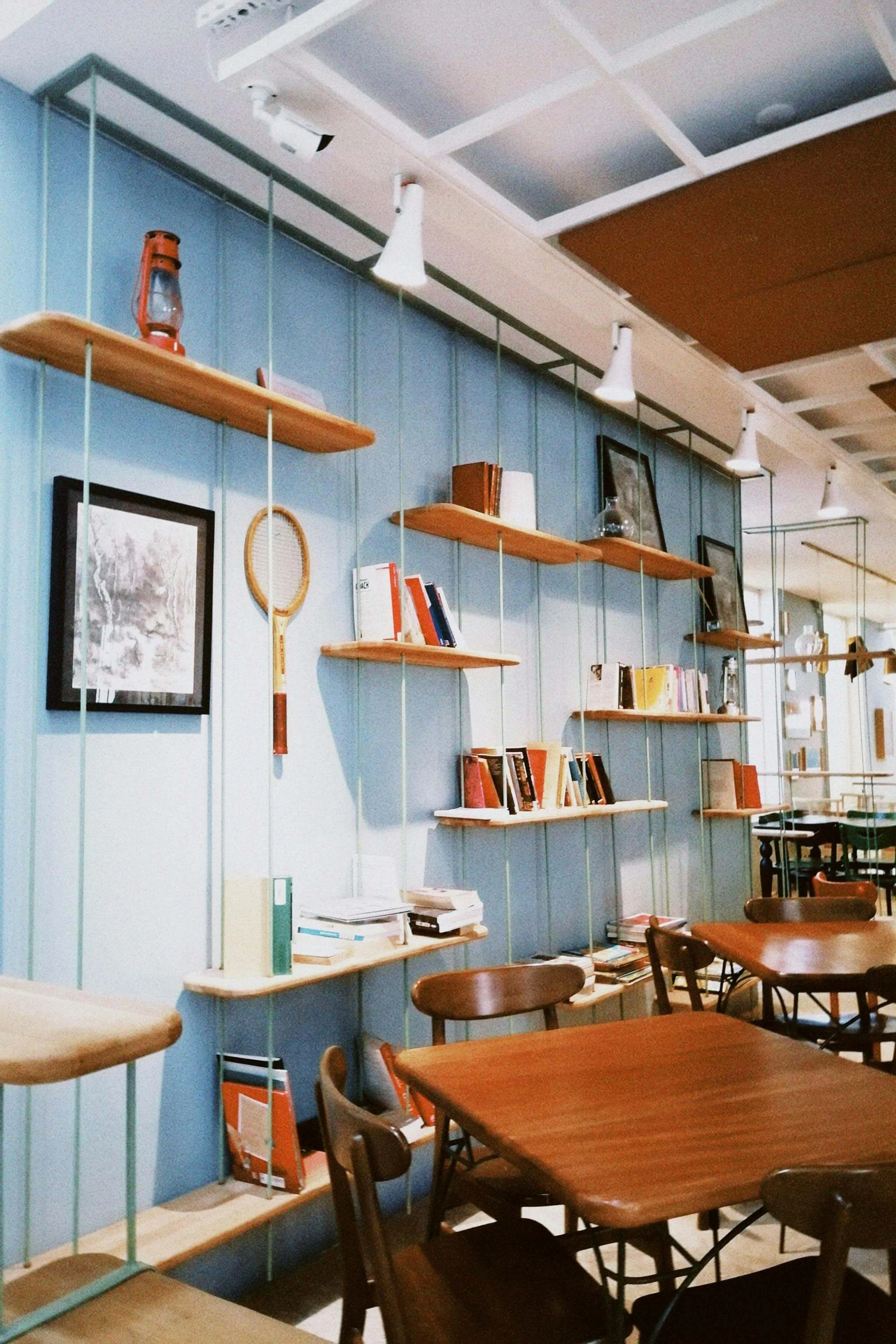 Une section d'une bibliothèque ou d'un café avec des étagères en bois fixées au mur bleu, contenant des livres, des cadres et des objets décoratifs. La zone comprend également des tables en bois et des chaises en bois.