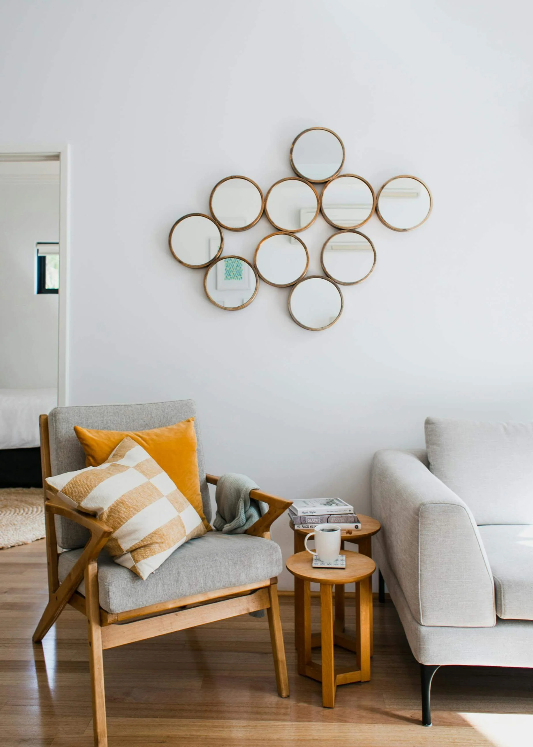 Intérieur d'un salon avec un fauteuil en bois et tissu gris, une table d'appoint en bois avec une tasse, des livres et un ordinateur portable, un canapé gris clair, et un mur blanc avec un ensemble de miroirs ronds encadrés en bois.