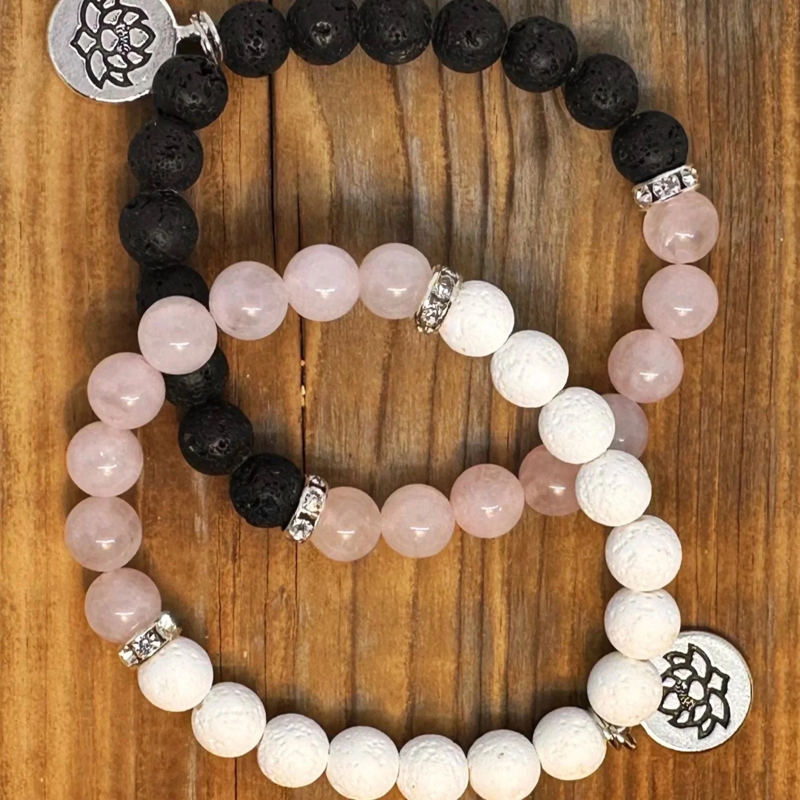 Two beaded bracelets, one black with lava stone beads and the other pink and white with textured beads, adorned with silver accents and charms, resting on a wooden surface.