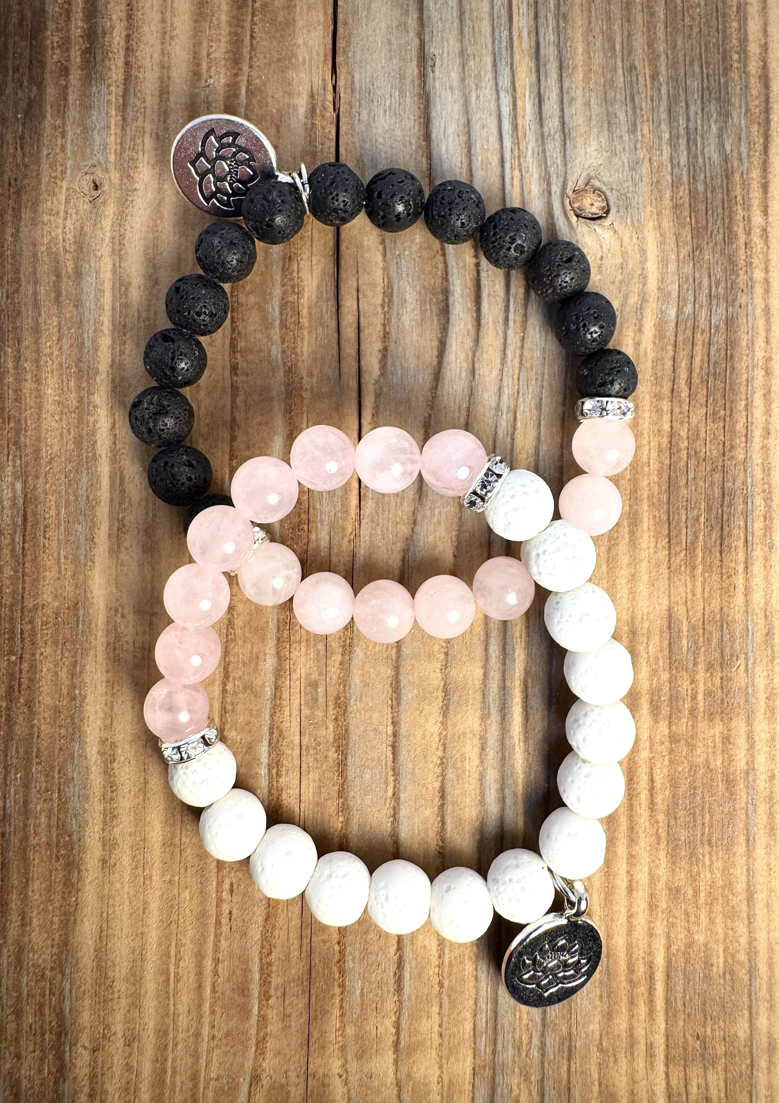 Two beaded bracelets, one black with lava stone beads and the other pink and white with textured beads, adorned with silver accents and charms, resting on a wooden surface.