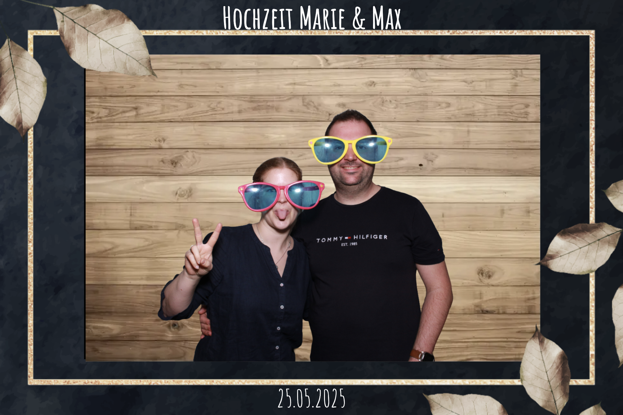 Ein Paar auf einer Hochzeitsfeier trägt große Sonnenbrillen, das Mädchen macht ein Peace-Zeichen und sie stehen vor einem Holz hintergrund, mit dem Text "Hochzeit Marie & Max" und dem Datum 25.05.2025.