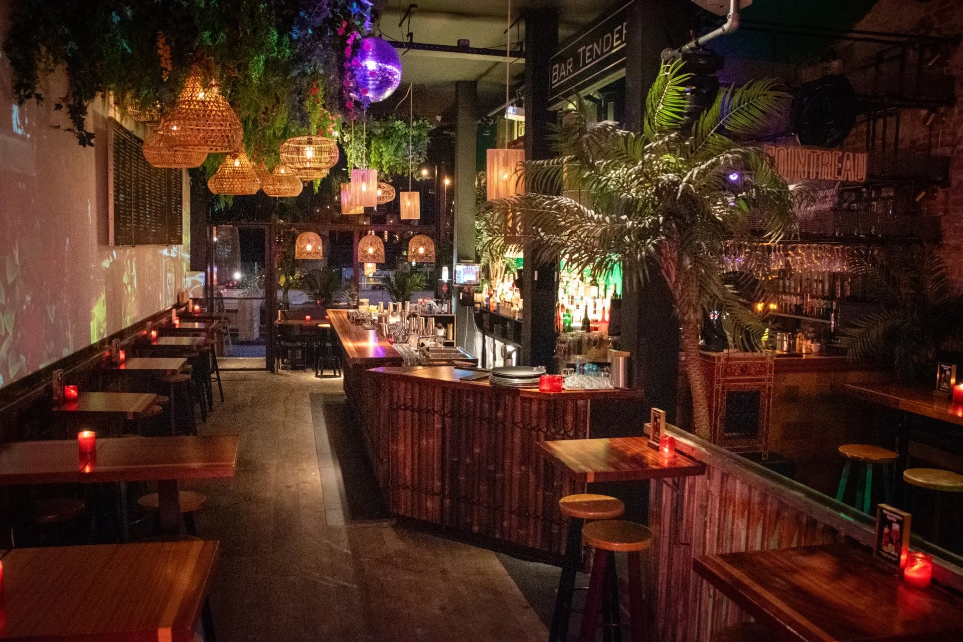 Binnenkant van een bar met houten tafeltjes, stoelen, sfeerverlichting, planten en een bar met diverse drankflessen.