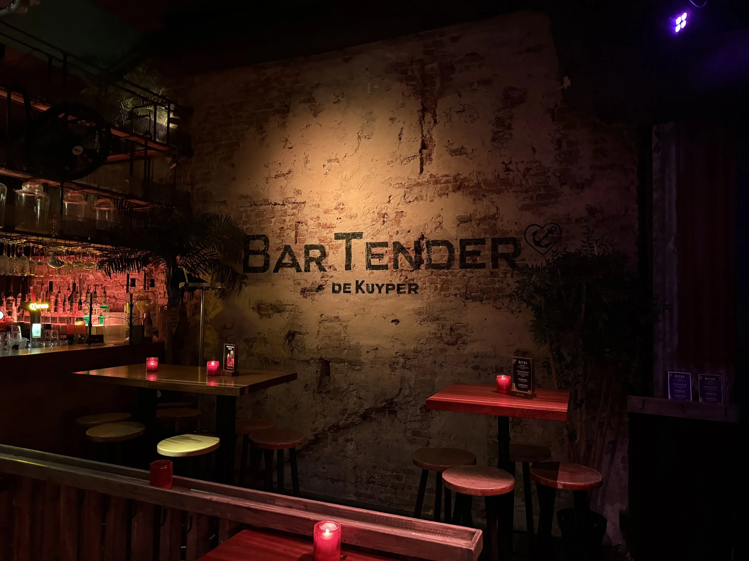 Interieur van een bar met een ruige bakstenen muur, gedimde verlichting, rode kaarsen op de tafels, en een muur met de tekst 'Bar TENDER de Kuyper' en een tekening van een hart met een vork.