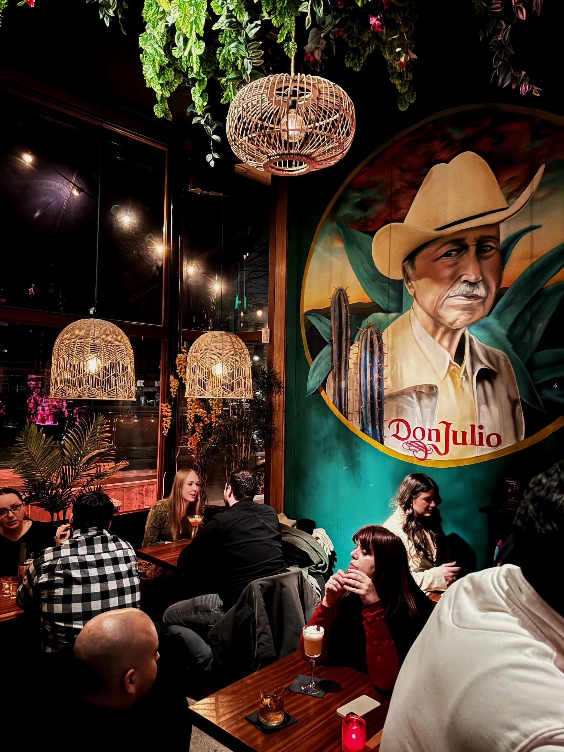 Een drukke restaurantruimte met mensen die genieten van drankjes, grote groene planten, hangende lampen, en een grote mural van een man met een cowboyhoed en de tekst 'Don Julio' op de muur.