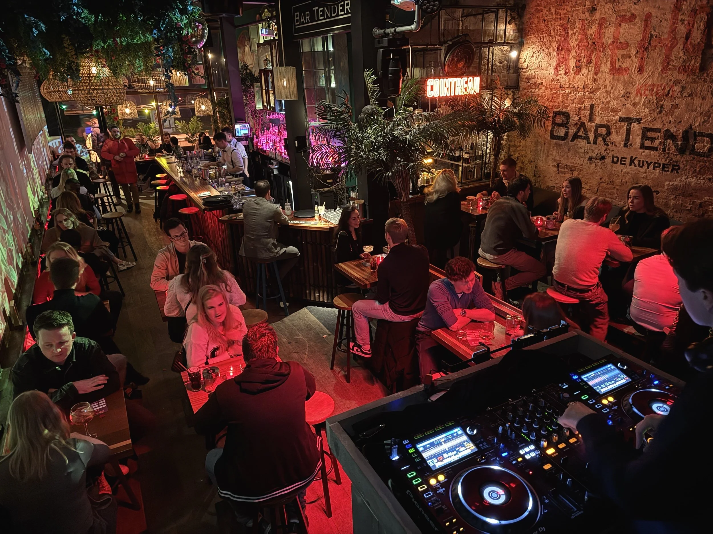 Een drukke bar met veel mensen die aan tafels zitten en genieten van drankjes, een DJ staat te draaien achtereenvolgens in de rechterhoek van de afbeelding en er hangen neonborden en planten in de ruimte.