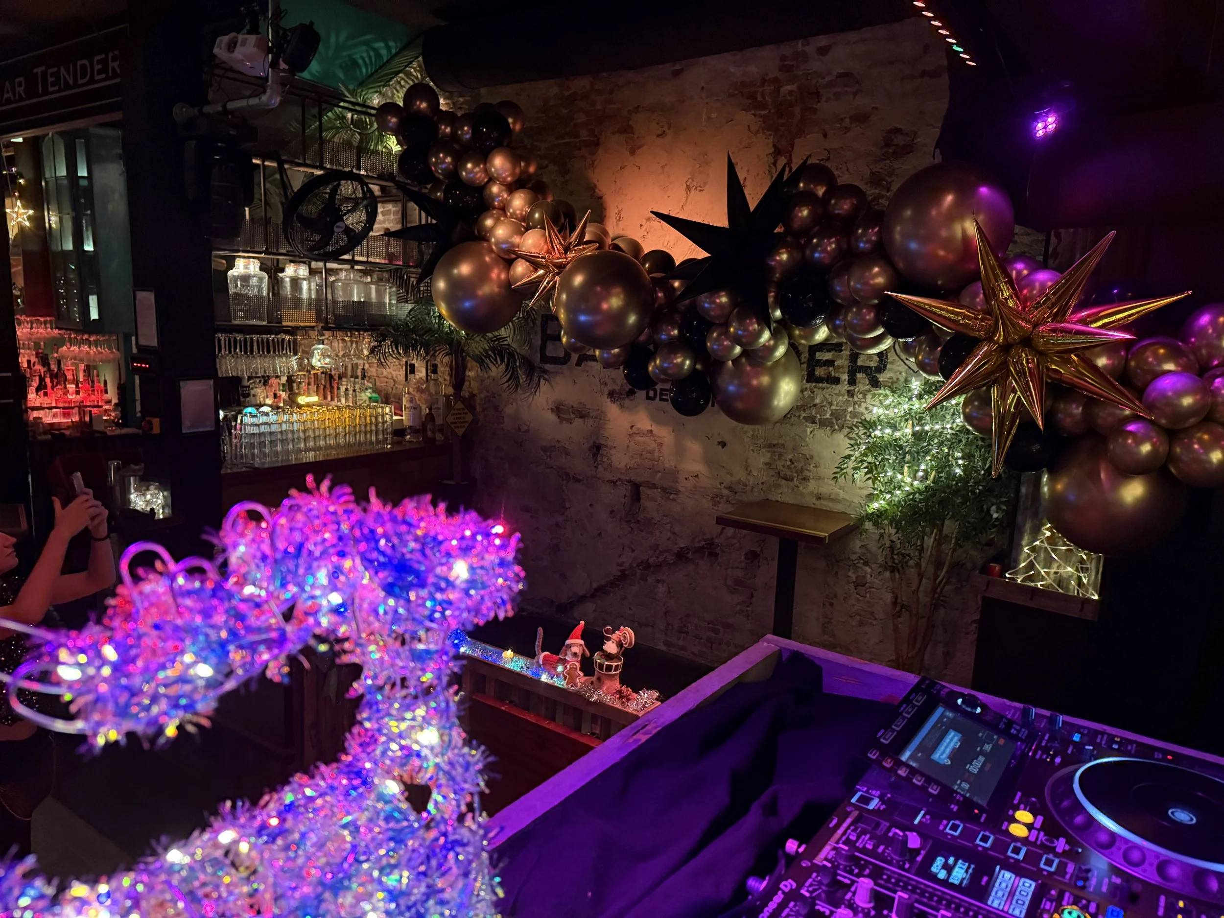 Feestdecoratie met ballonnen en sterren, feestverlichting, een bar met drankjes, en een DJ-setup met een mengpaneel en lichteffecten.