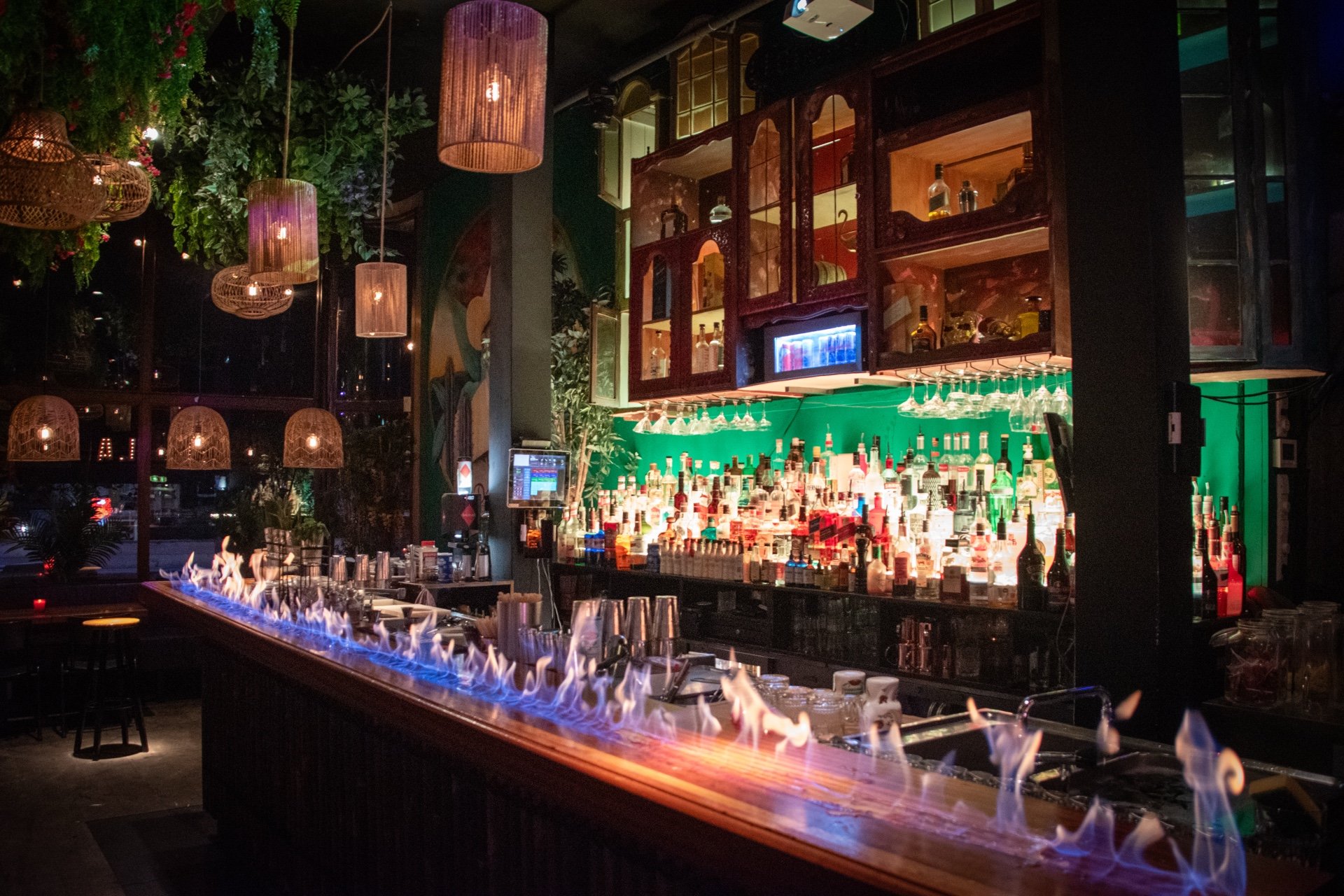 Een bar met talloze flessen drank en een lange bartafel met vlammen eronder, omringd door hangende lampen en decoraties.