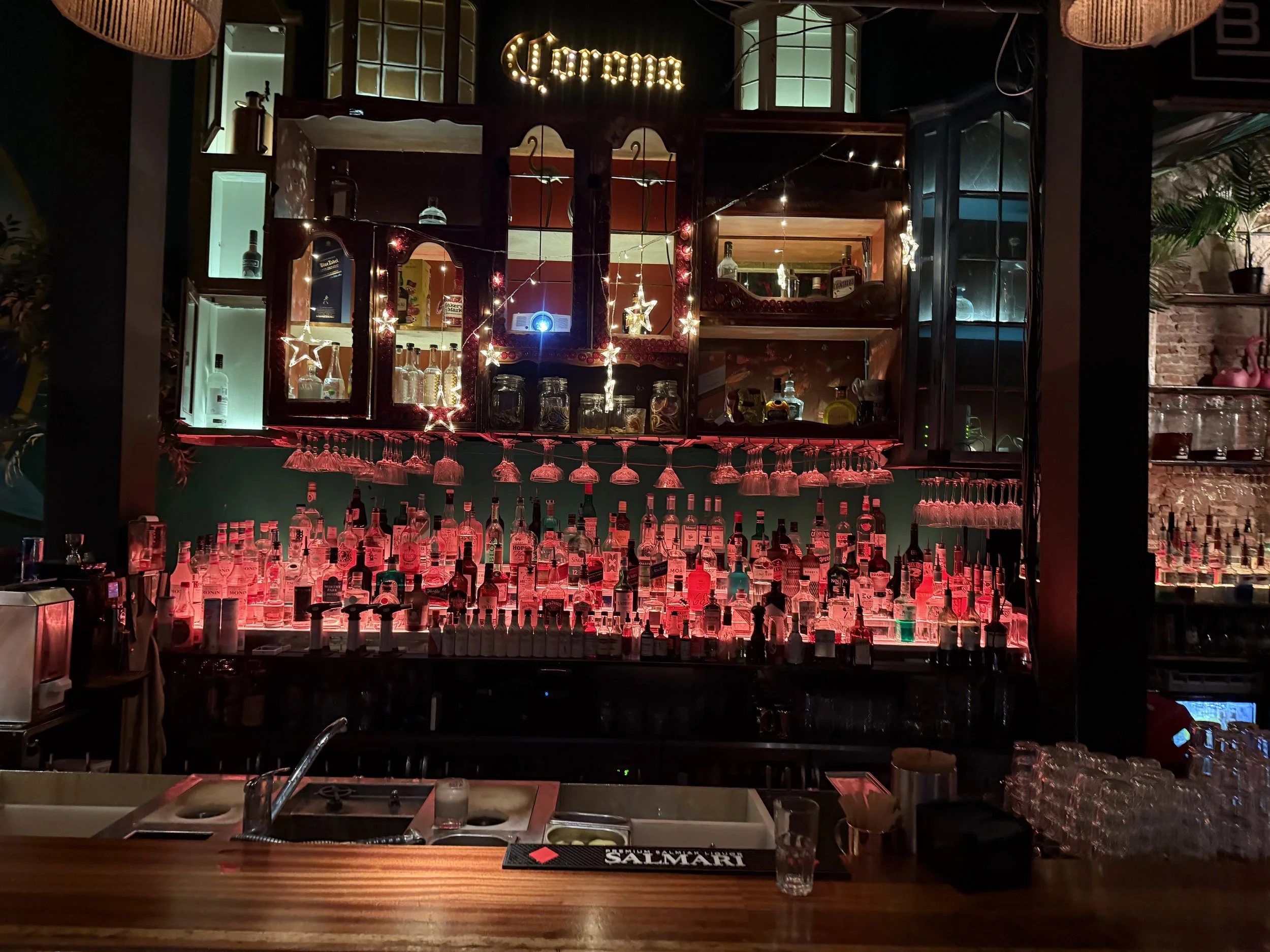 Een bar met een houten aanrecht, op de achtergrond een muur met glazen deurtjes en een verlichte signaal met de naam 'Corona'. Er hangt decoraties met sterren en lampjes, en er staan diverse alcoholflessen en glazen op de bar en daaronder.
