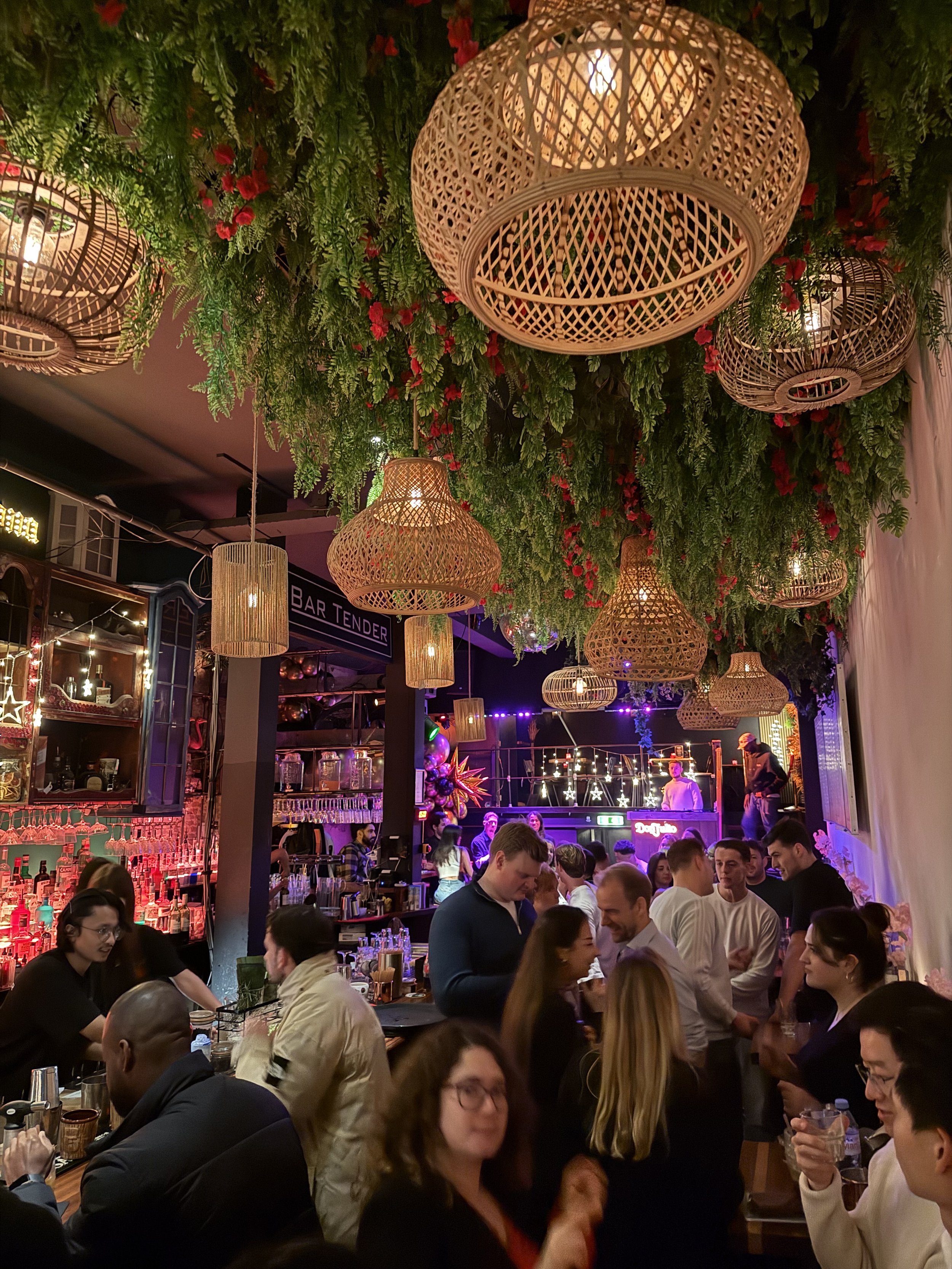 Een drukke bar of restaurant met mensen die aan de bar en tafels zitten. Het plafond is versierd met grote, ronde, mandachtige lichtlampen en groen blad met rode bloemen hangt van het plafond naar beneden. Men geniet van drankjes en gesprekken.
