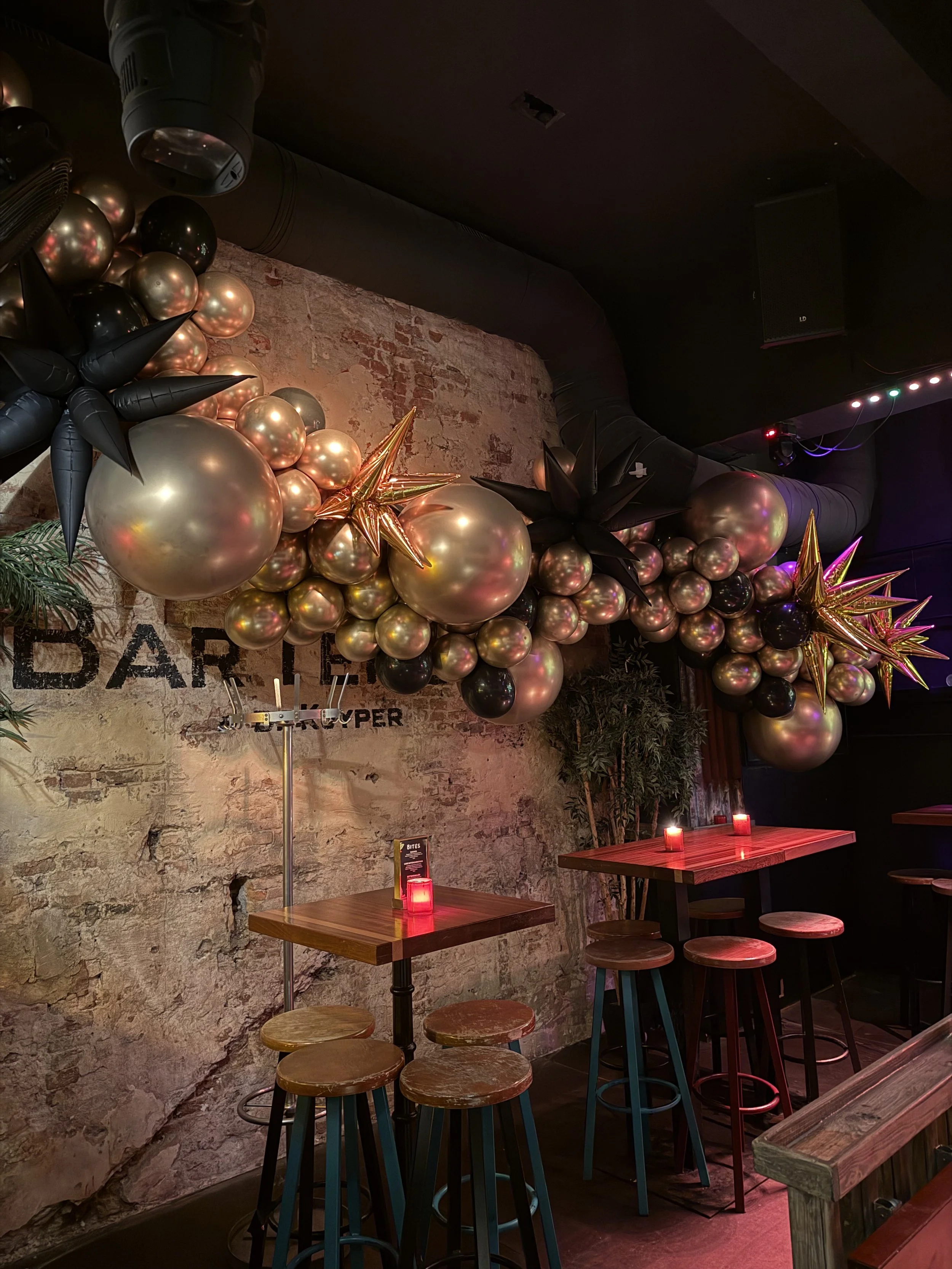 Binnenkant van een bar met een decoratie van goud en zwarte ballonnen en gouden sterren, kleine tafeltjes met rode lichten, houten krukken en een rotsmuur.