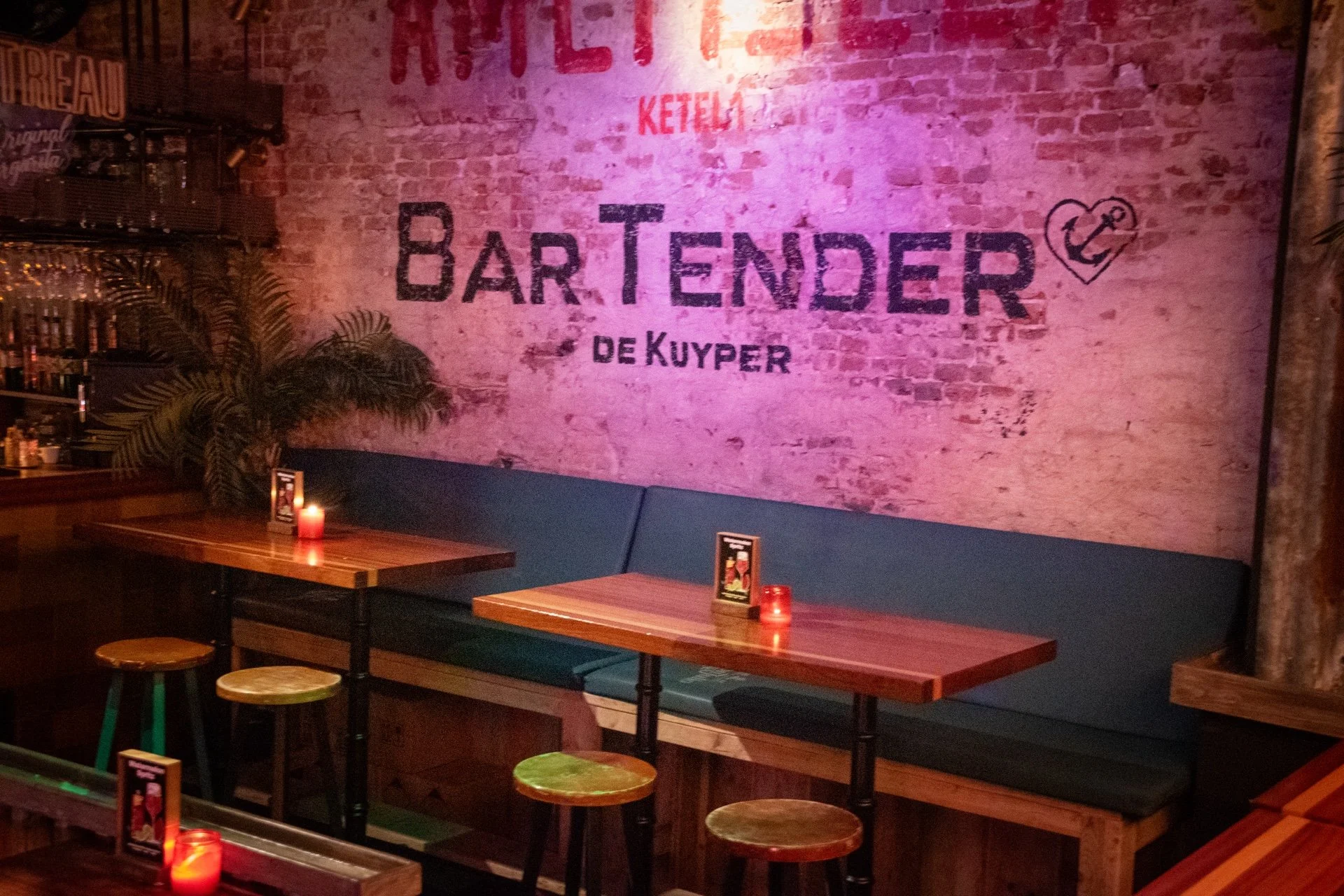 Binnenkant van een bar met een lage bank tegen een bakstenen muur, die beschilderd is met de woorden 'Bar Tender de Kuyper' en een hart met anker. Er staan tafels en barkrukken, met kaarslicht op de tafels.