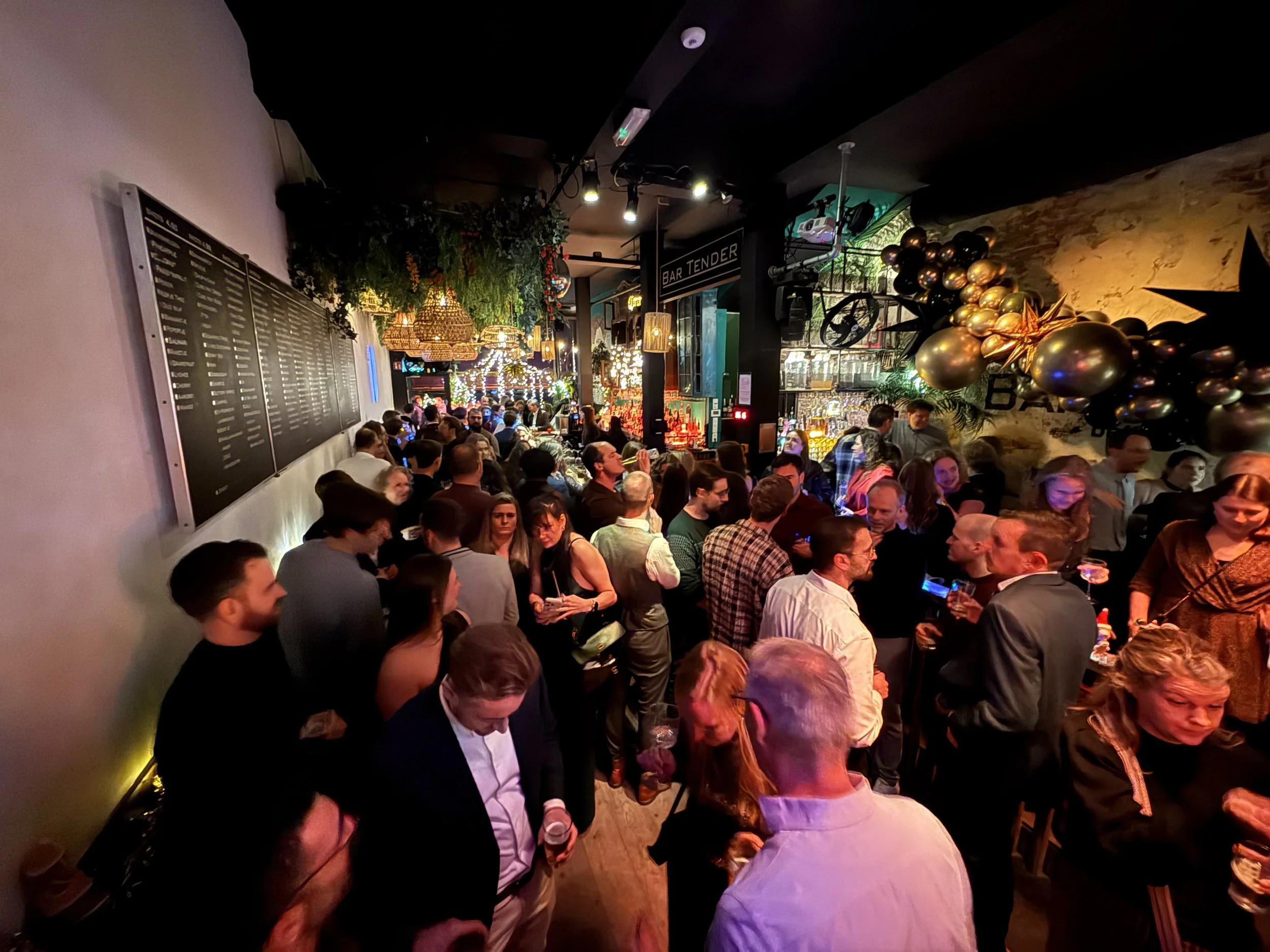 Een drukke bar of club met veel mensen die genieten en praten. Decoraties met ballonnen en verlichting, een menu aan de muur hangen en een bar op de achtergrond.