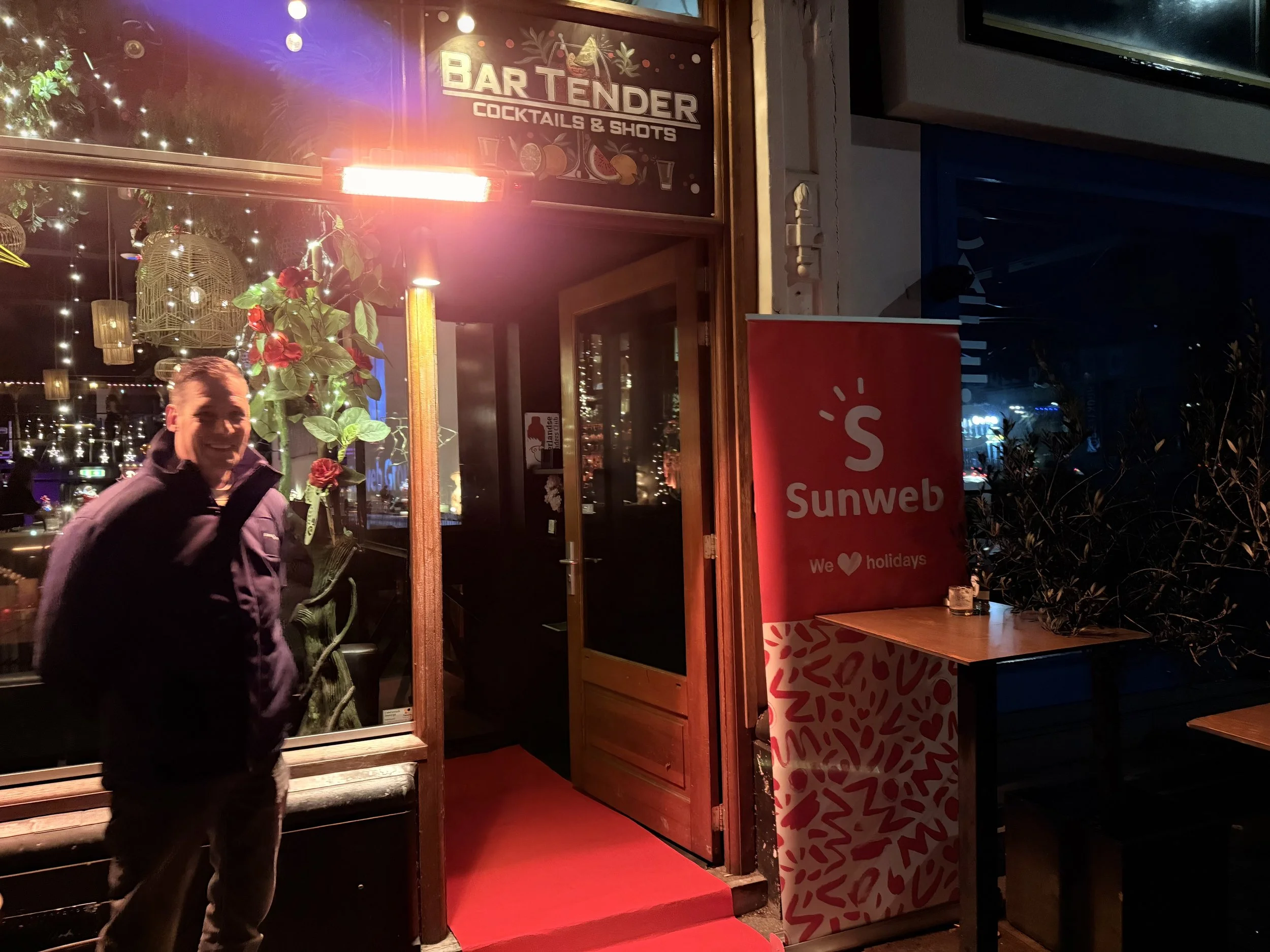 Man die de deur van een bar opent, met een grote rode loper en een bordje met de tekst 'Bar Tender Cocktails & Shots'.'}】'avenir.  In english: 