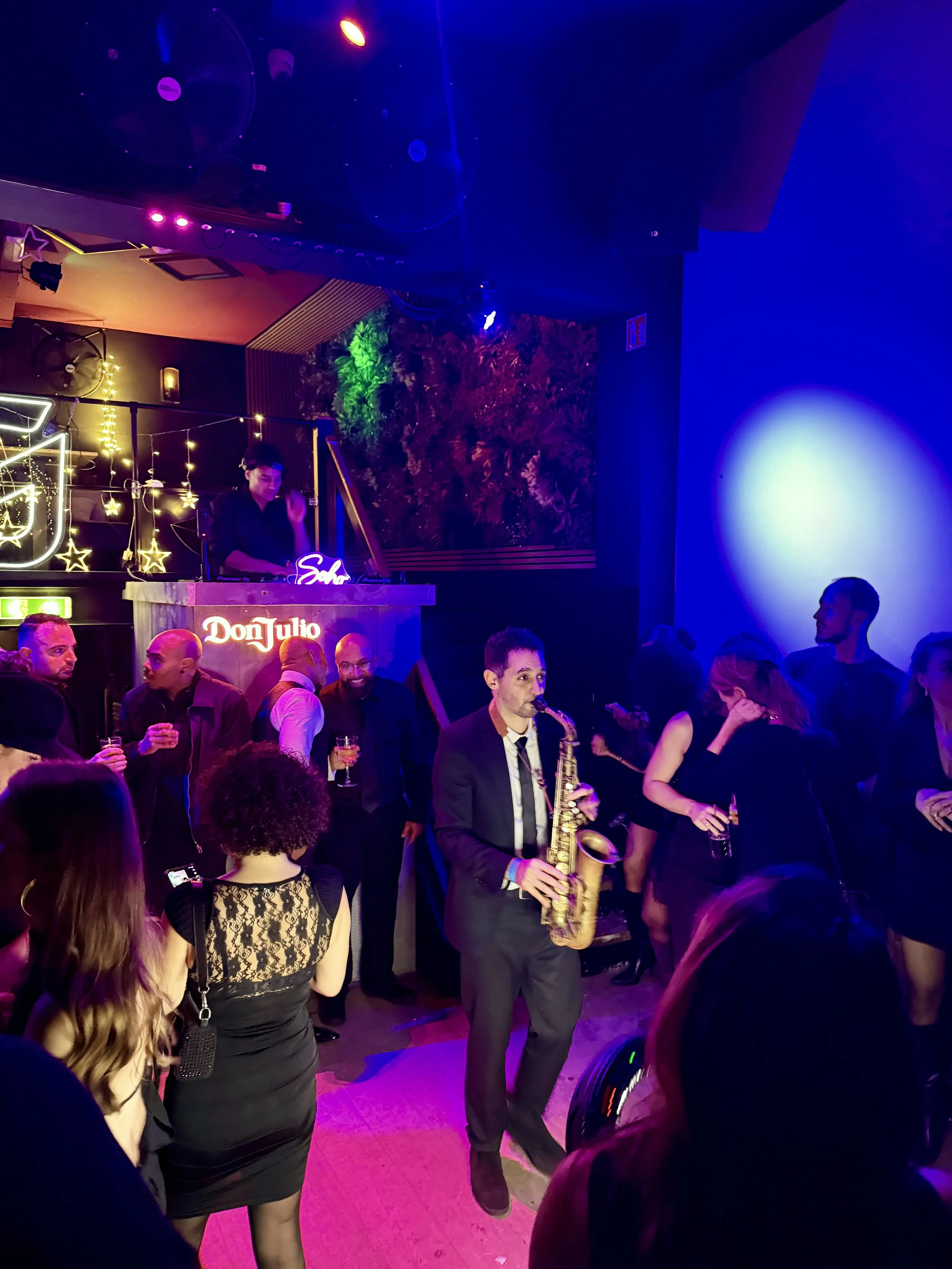 Mensen dansen en genieten van een live jazz band in een club met kleurrijk licht, een saxofonist in het midden draagt een pak en een das.