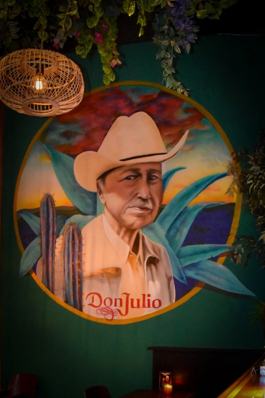 Muurkunst met een portret van Don Julio, een man met een cowboyhoed en een beige jas, omgeven door cactussen en agaveplanten, met een kleurrijke zonsondergang op de achtergrond.