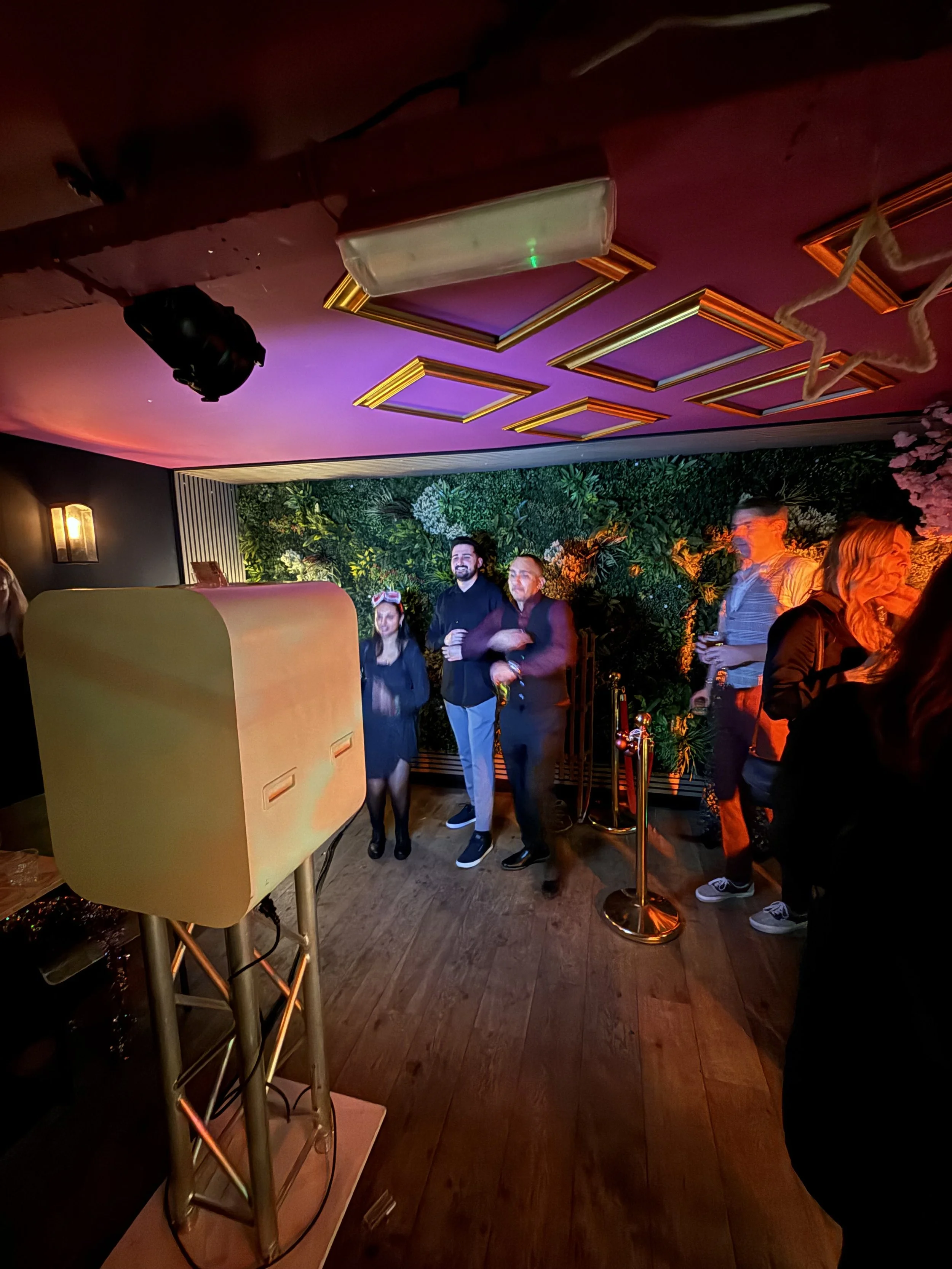 Binnen bij een feest of evenement, mensen staan en praten, er hangt kleurrijk decor, en er zijn stopcontacten en een volledige muur met groene planten.