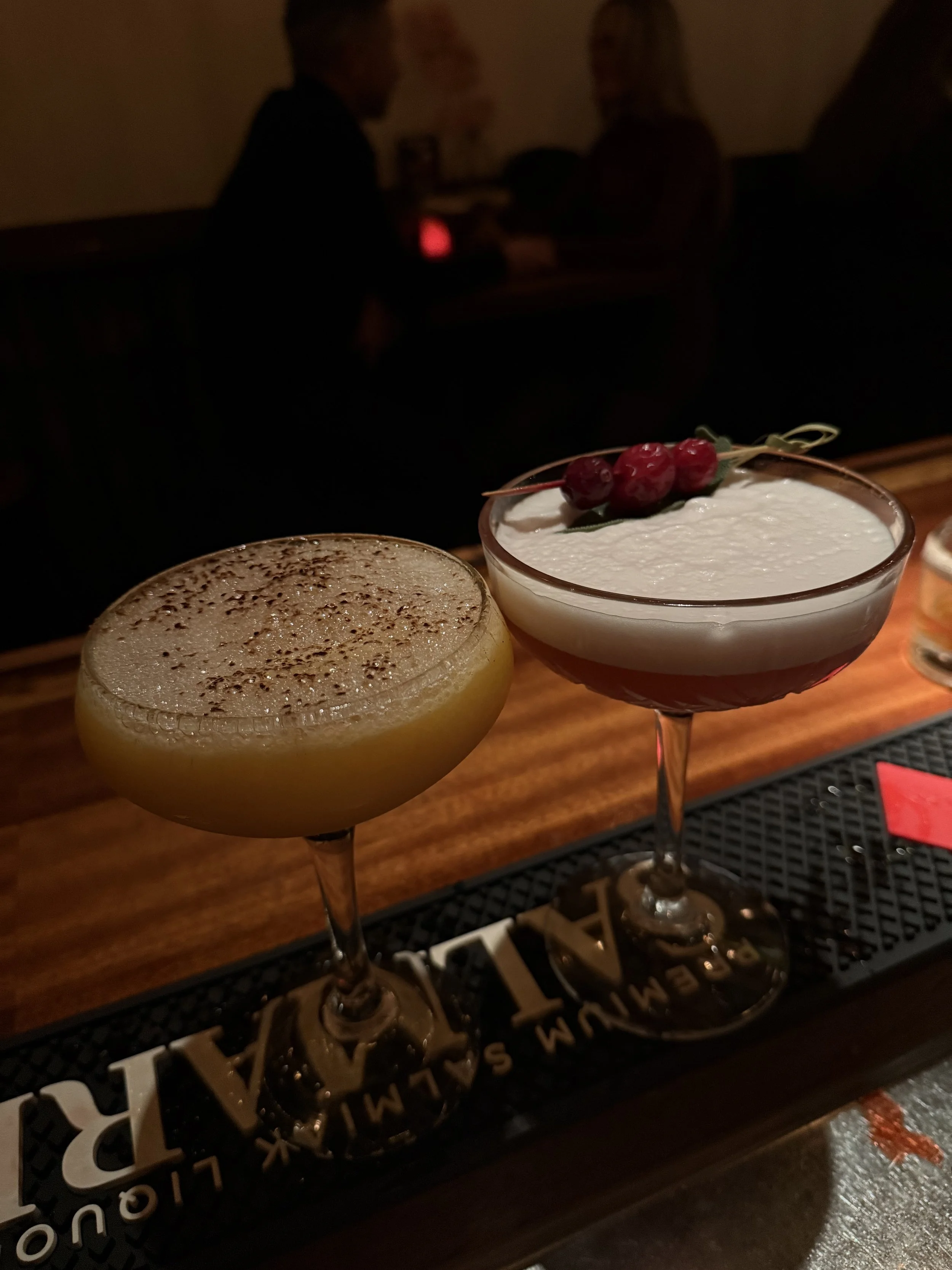 Twee cocktails op een donkere barkruk, een met schuim en cacaopoeder en de ander met room en kersen als garnering.